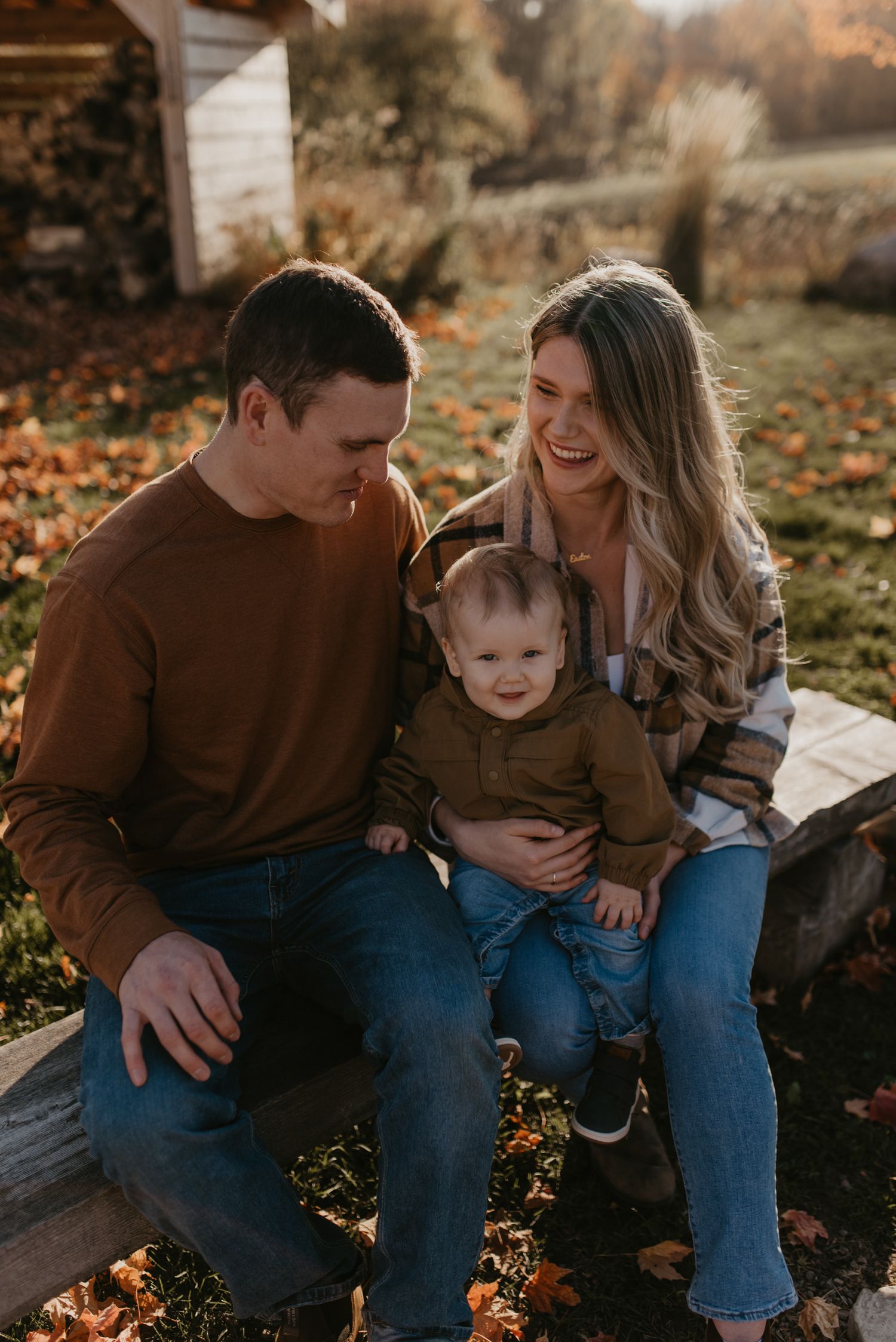 A happy young family with a baby sits outdoors on a fall day, surrounded by colorful autumn leaves.