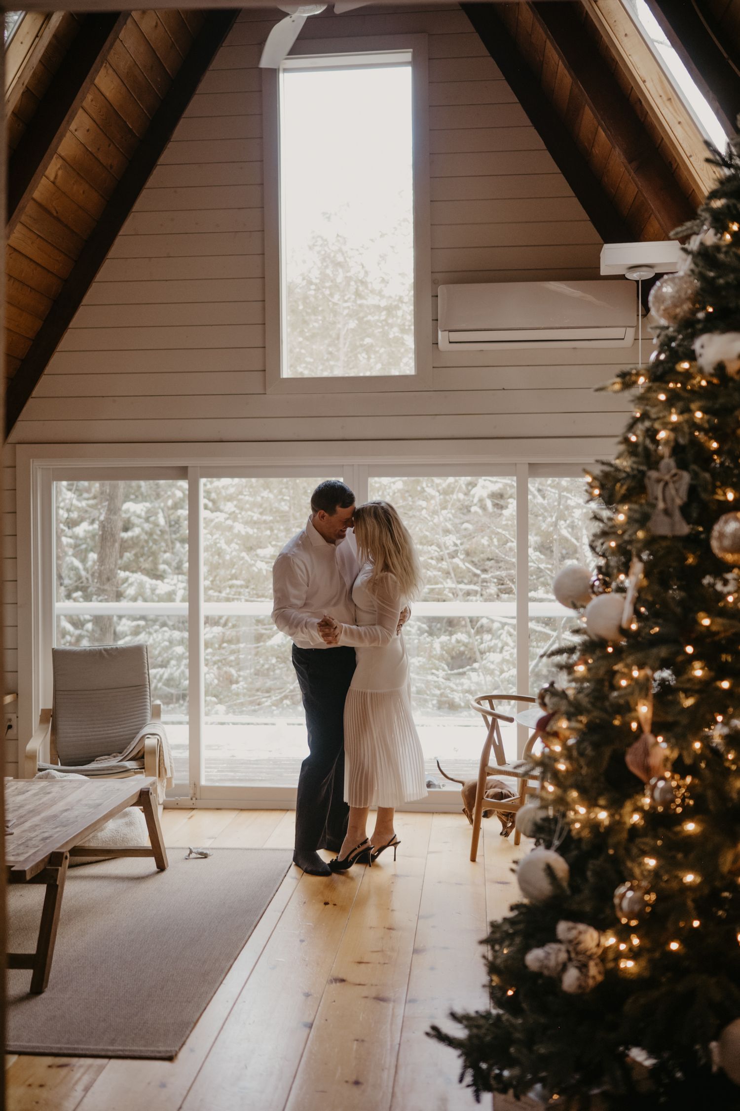 Romantic couple sharing a kiss near a decorated Christmas tree inside a bright A-frame cabin with snowy view.