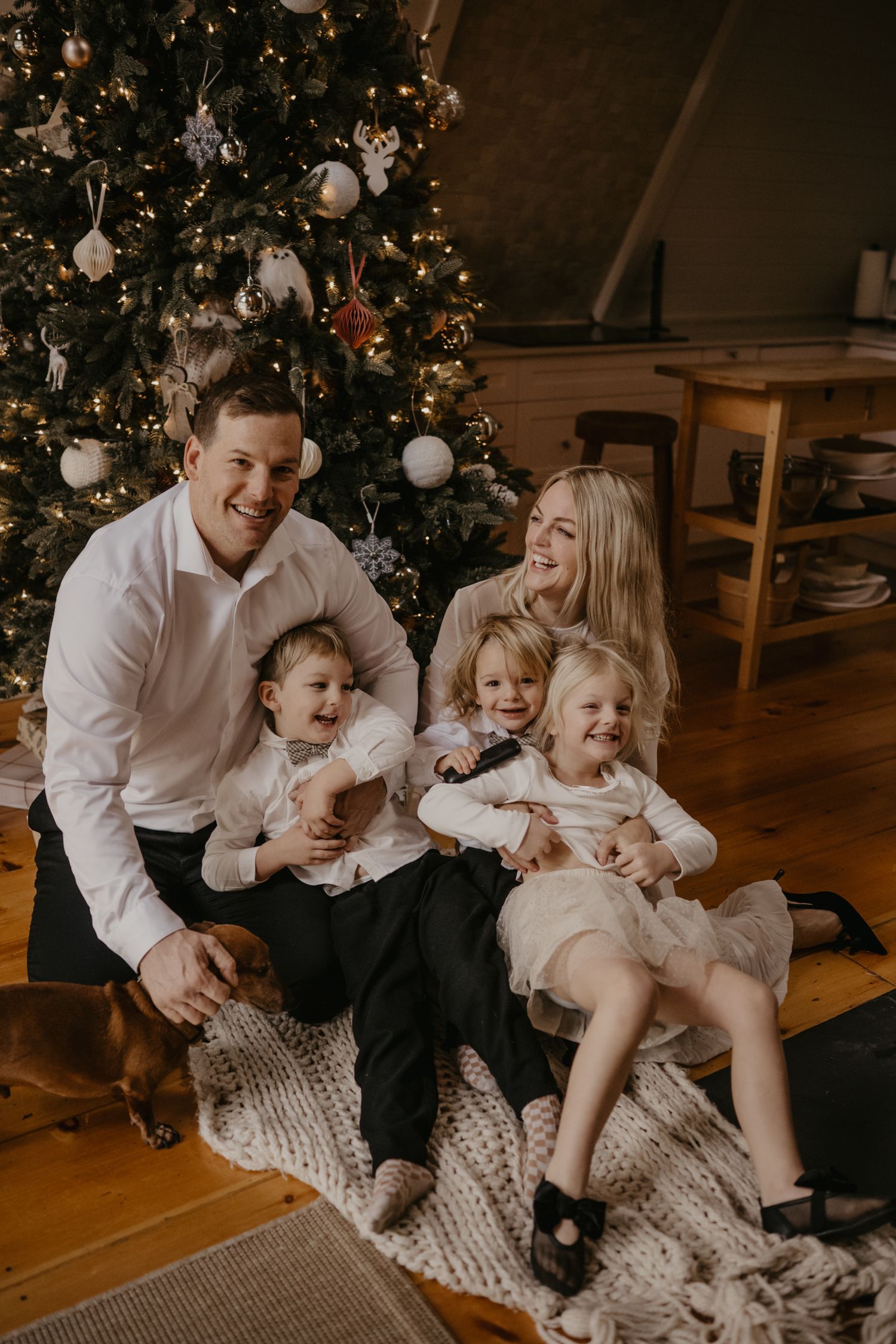Happy family with three young children sitting on a blanket in front of a decorated Christmas tree with a dachshund.
