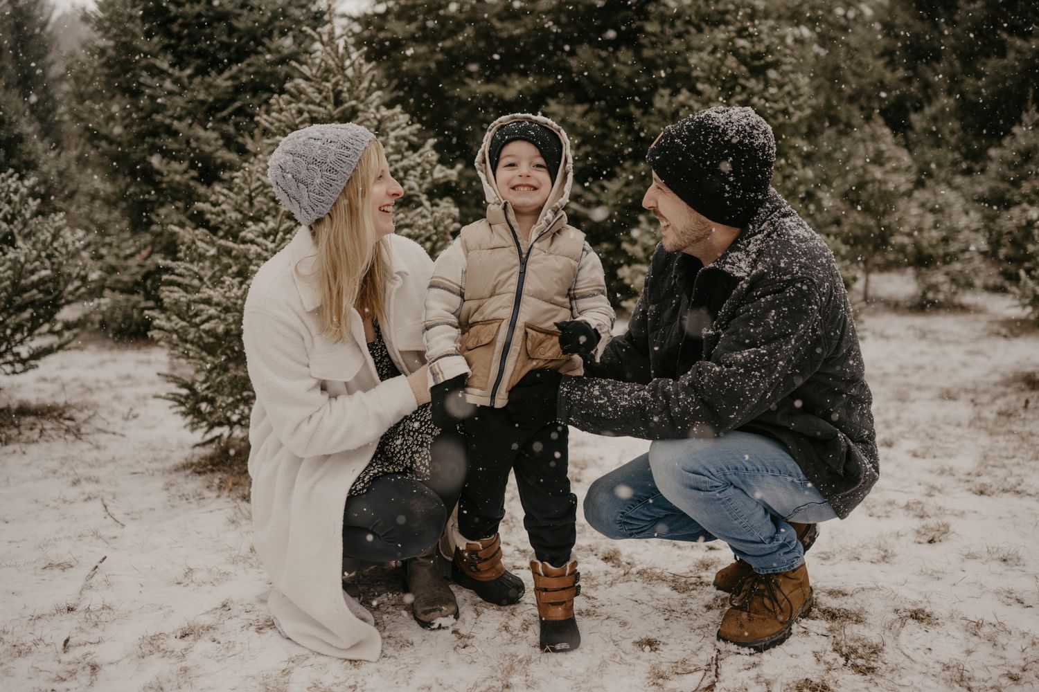 Family with young child playing in snow at a Christmas tree farm, parents crouching beside toddler in winter gear.