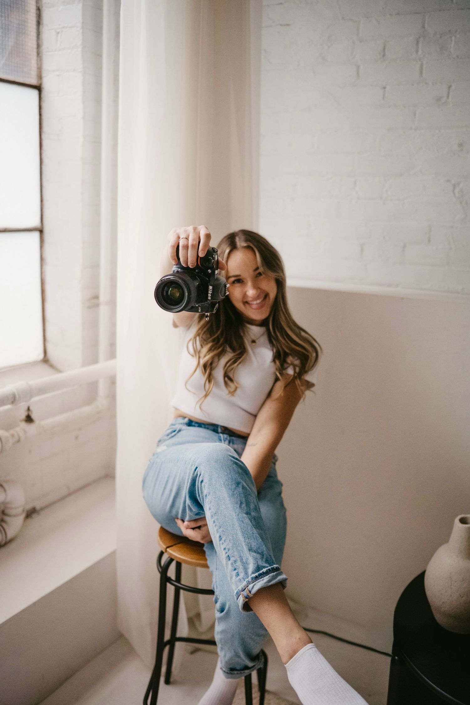 Smiling female photographer holding up DSLR camera while seated on stool in bright white studio.
