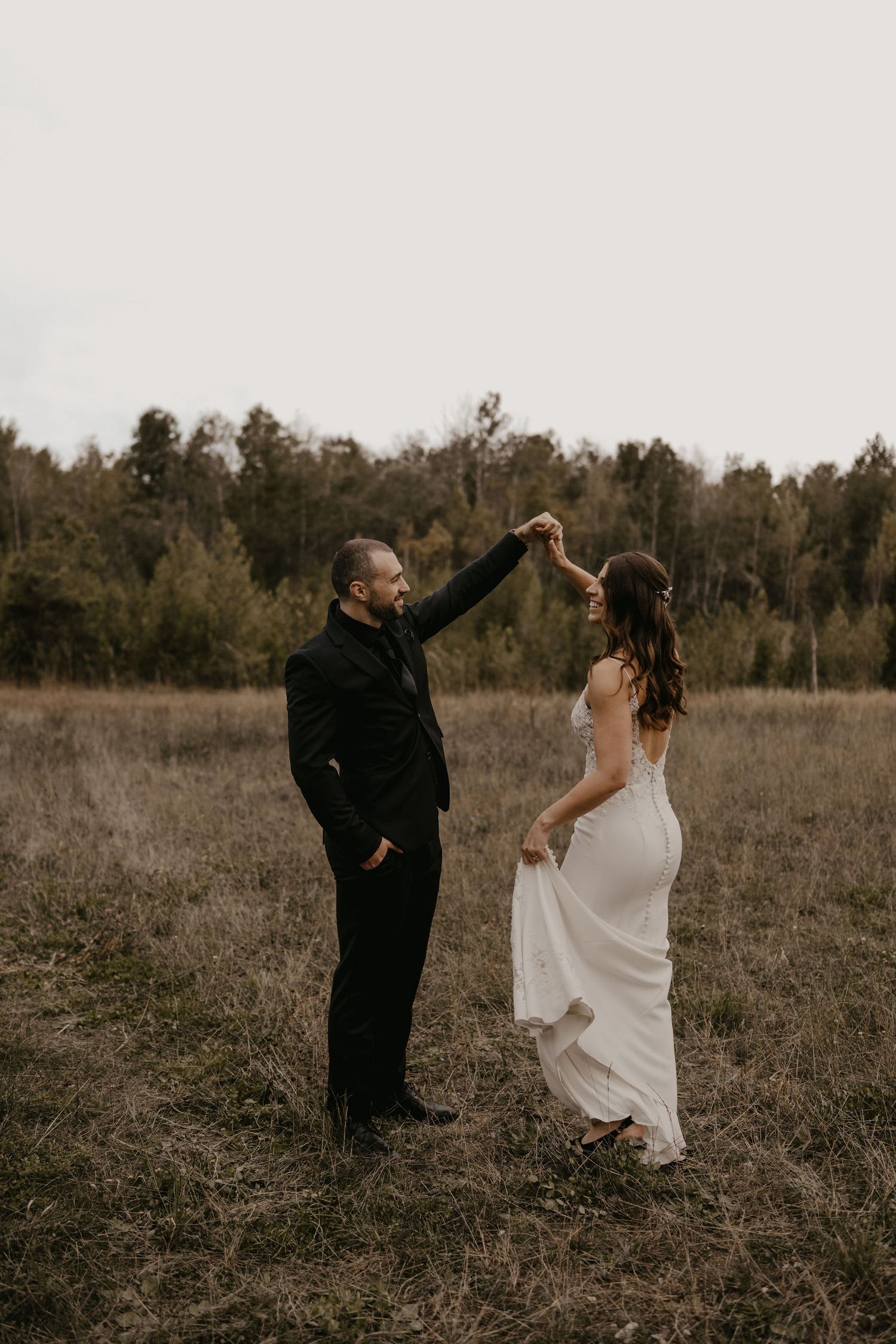 Groom twirls bride in white dress in an open autumn field with forest backdrop.