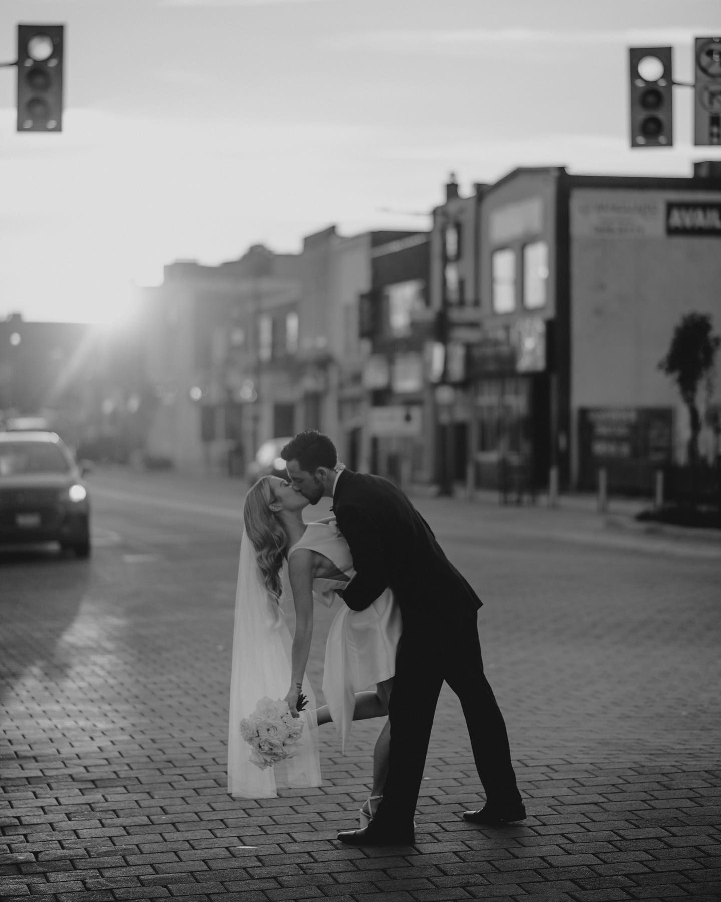 Black and white photo of bride and groom sharing a kiss on a city street at sunset.