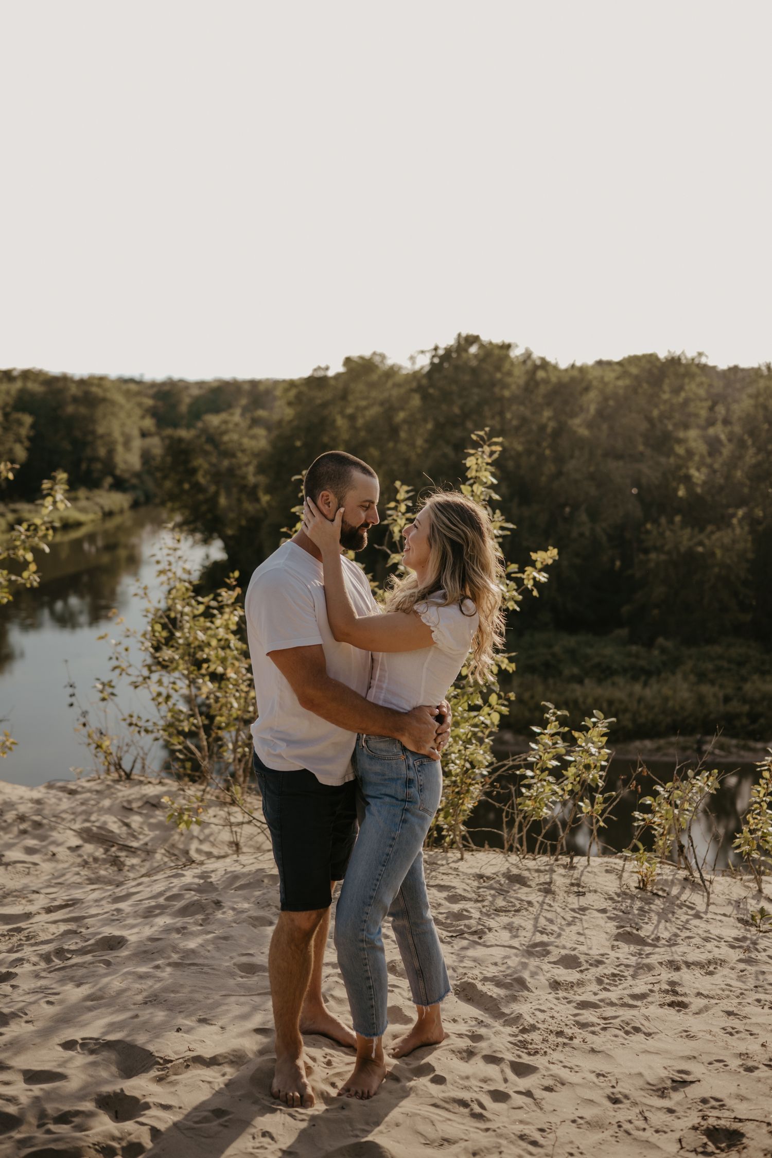 Couple embracing and gazing at each other on sandy dunes with river and green trees at sunset.