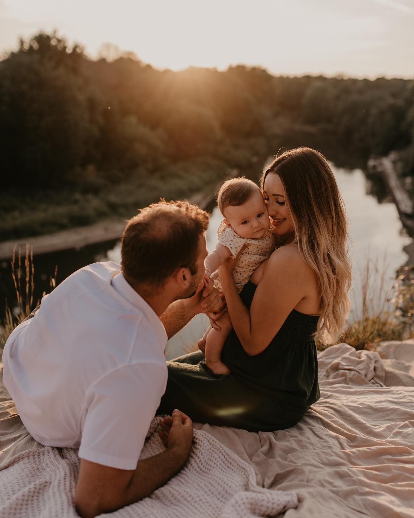 Family enjoying a golden sunset outdoors, parents lovingly holding their baby on a blanket by a river.