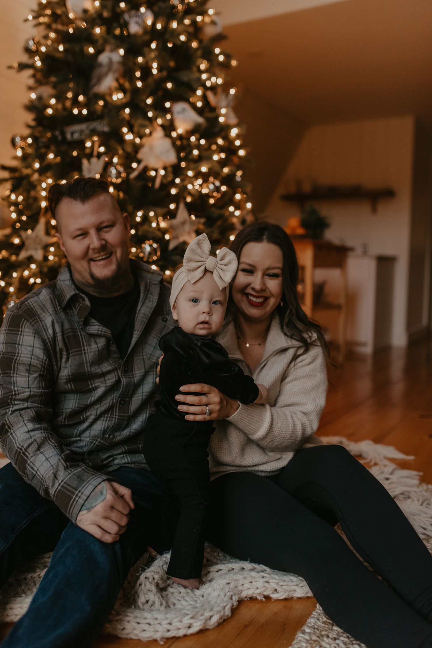A smiling family with a baby girl in a bow sits on a rug in front of a decorated Christmas tree.