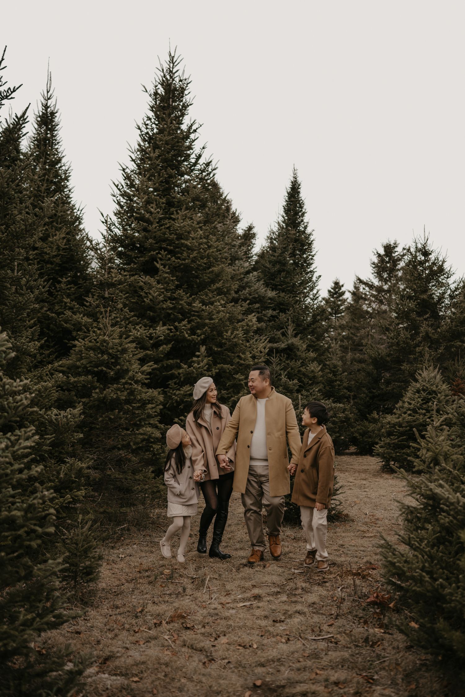 Family of four walking through a Christmas tree farm in coordinating neutral beige winter outfits.