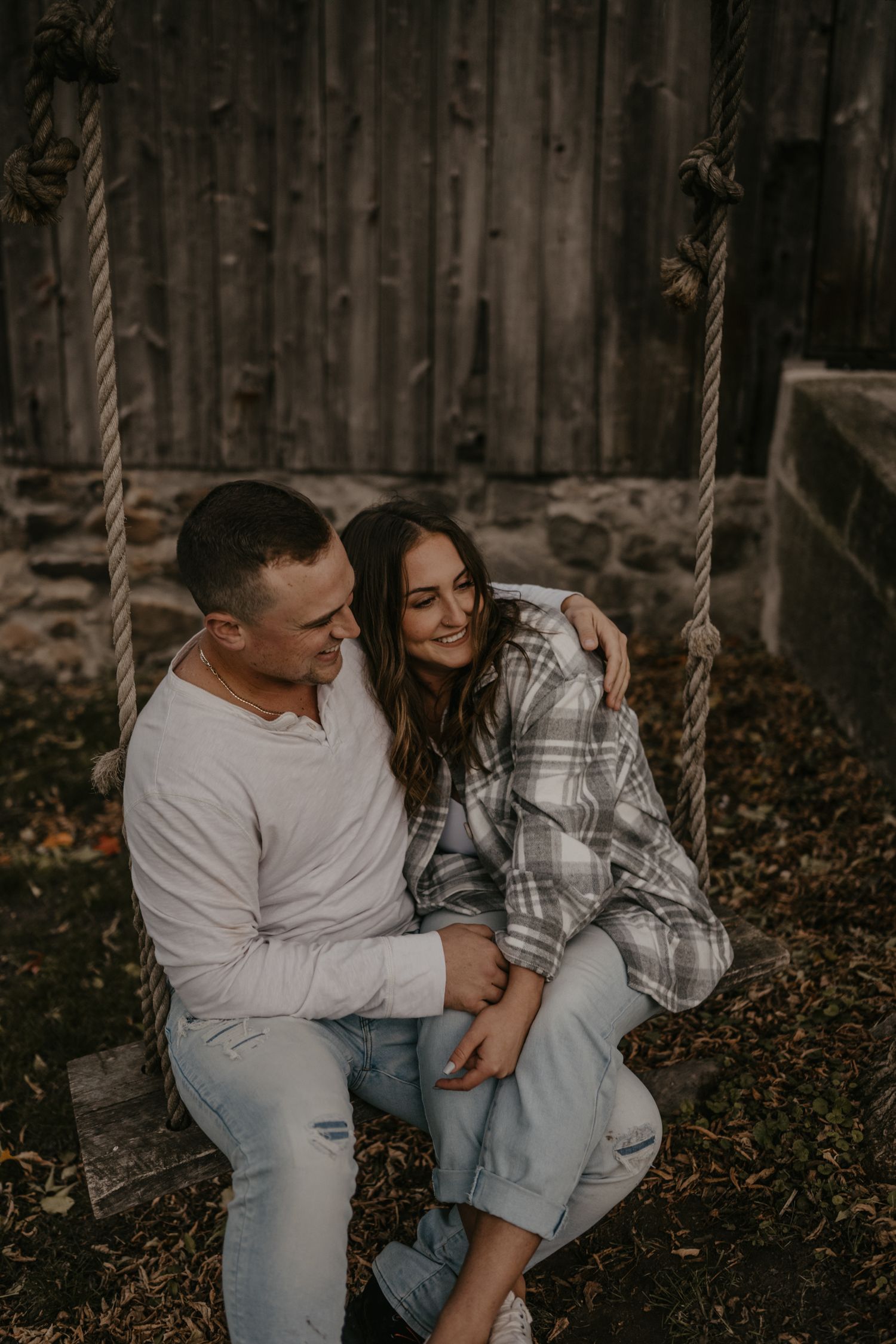 Couple sits closely on rustic rope swing, smiling together in warm autumn light.