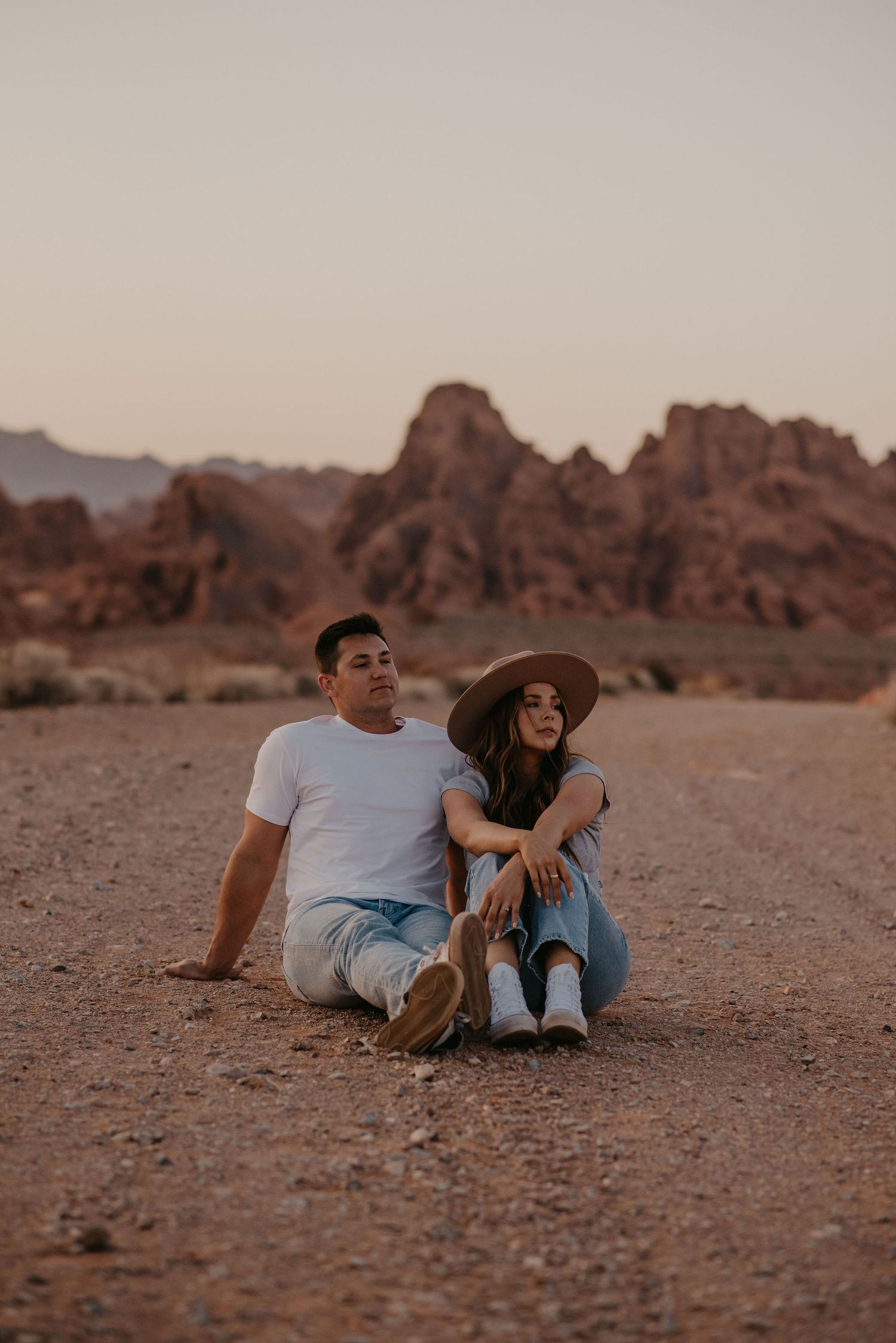 Couple sitting together on desert ground with red rock formations at sunset.