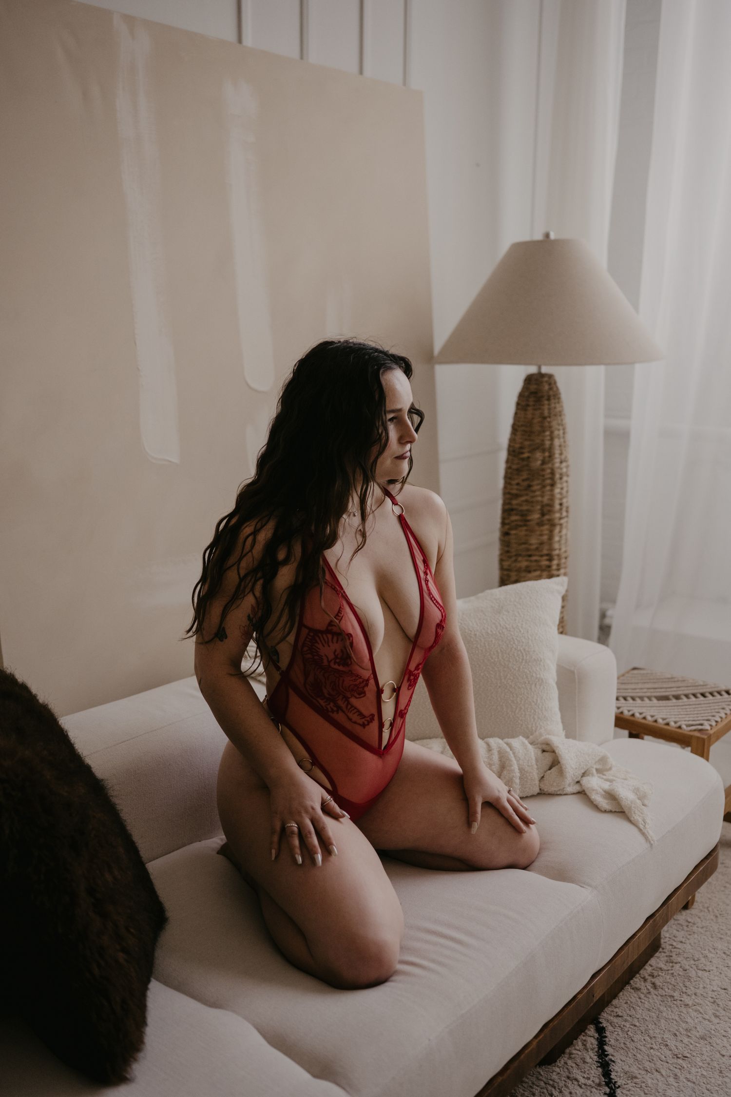 Woman in red lace bodysuit kneeling on a chaise lounge in a softly lit, minimalist room.