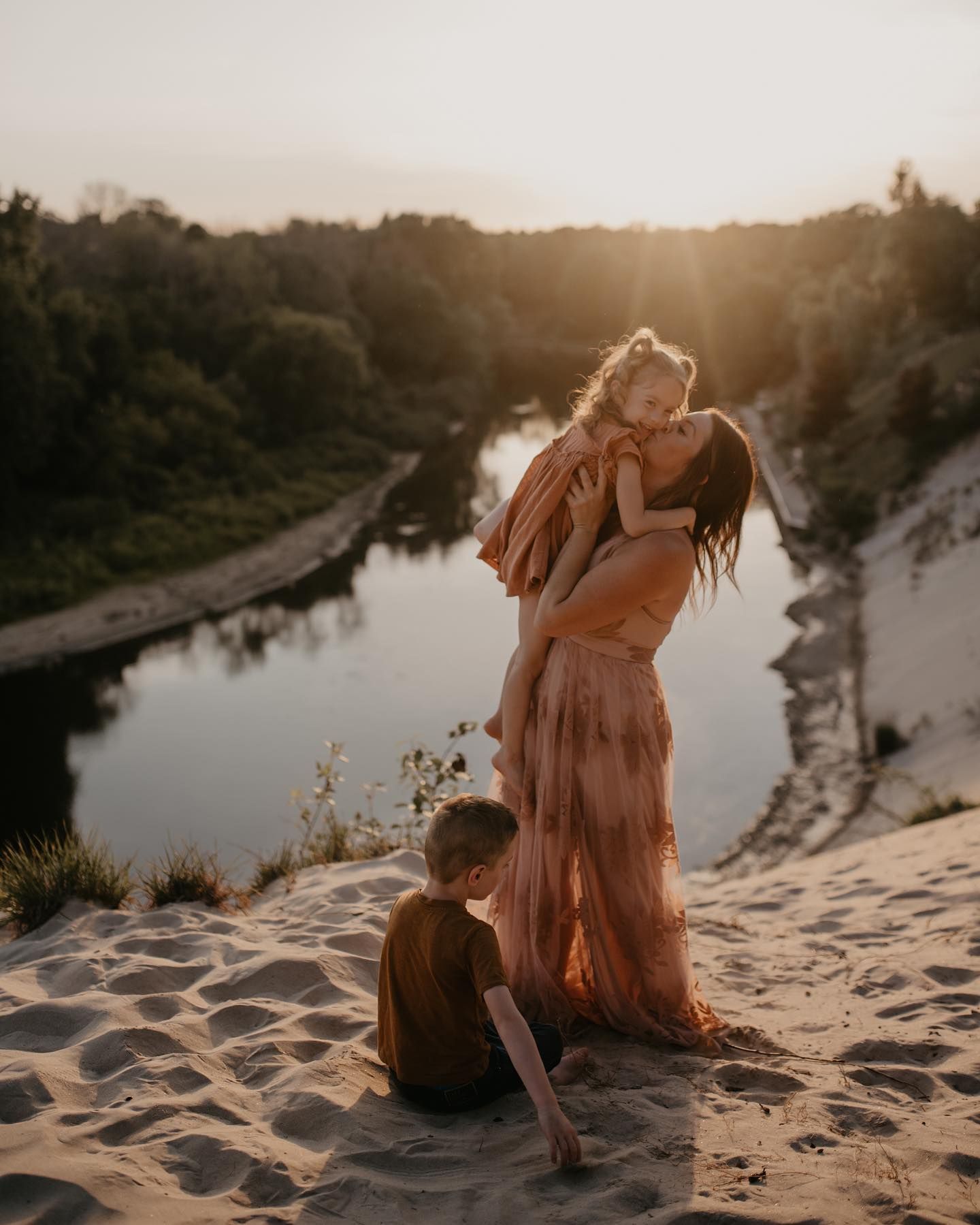Mother lifting child at golden hour on sandy cliff by river, with son sitting nearby in warm sunset light.
