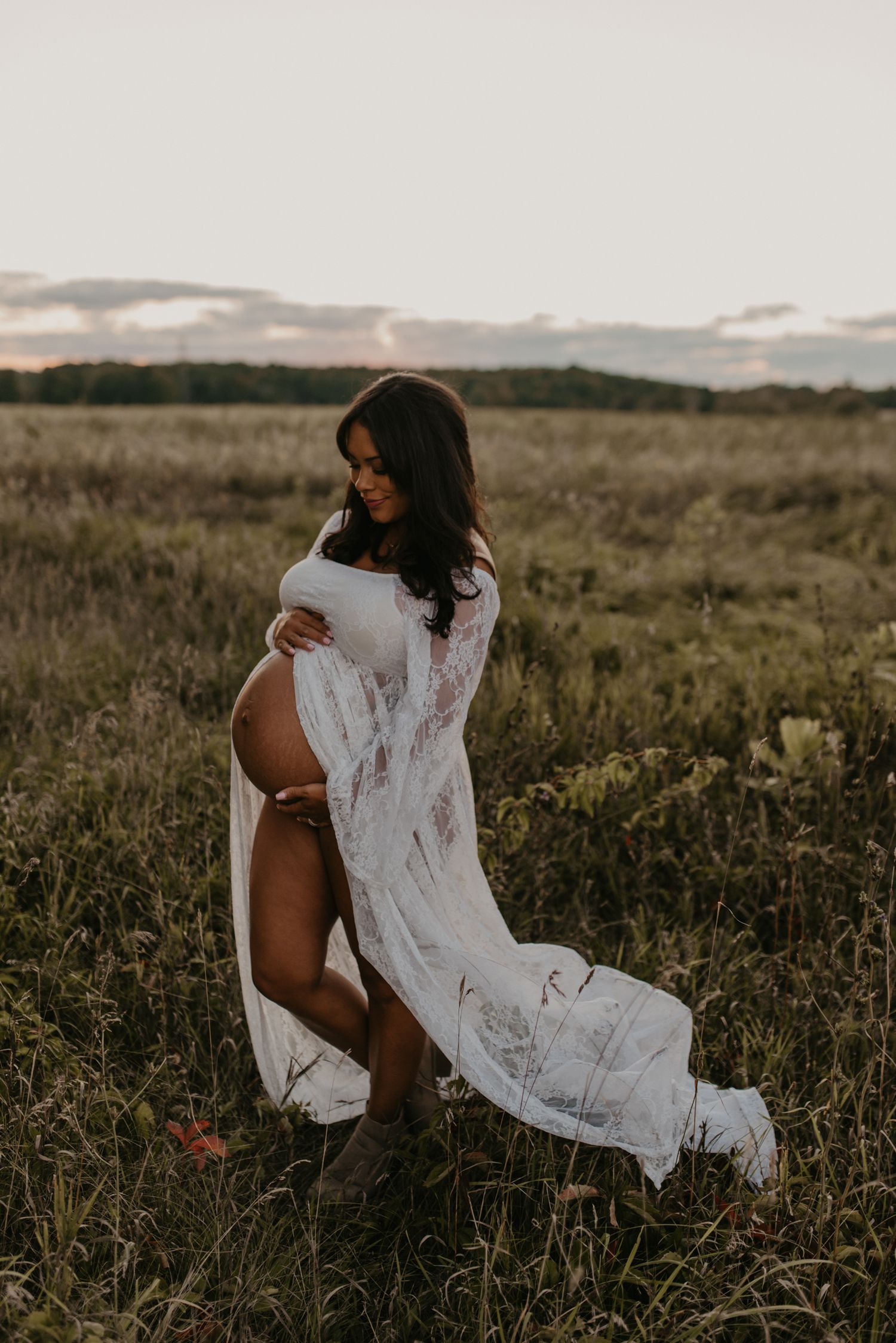 Pregnant woman in flowing white lace gown cradling her baby bump in a golden field at sunset.