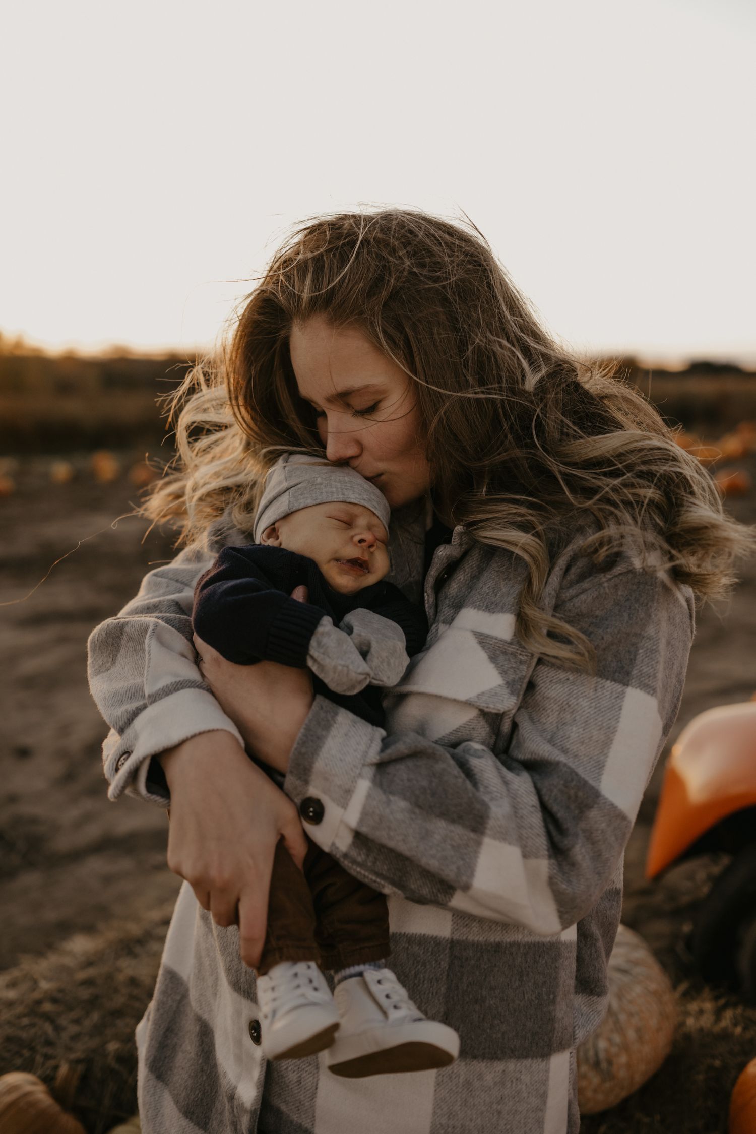 New mother tenderly kissing her sleeping newborn baby at sunset in a pumpkin patch.