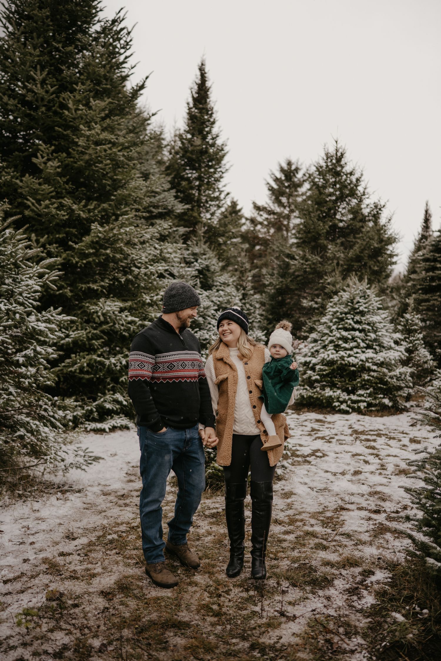 Family of three poses among snow-dusted evergreen trees at a Christmas tree farm in winter.