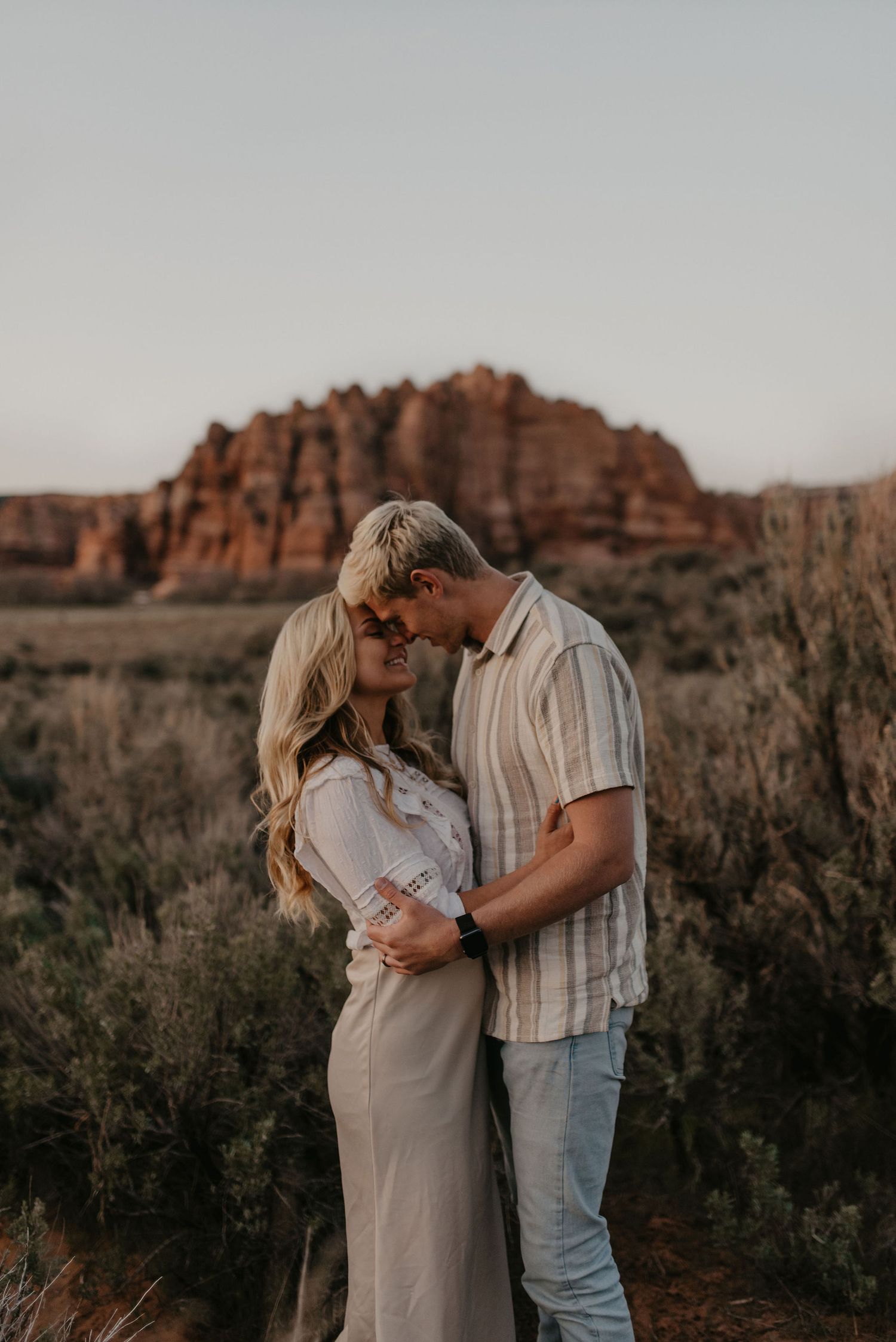 Couple embracing and smiling at each other near red rock formations at golden hour.