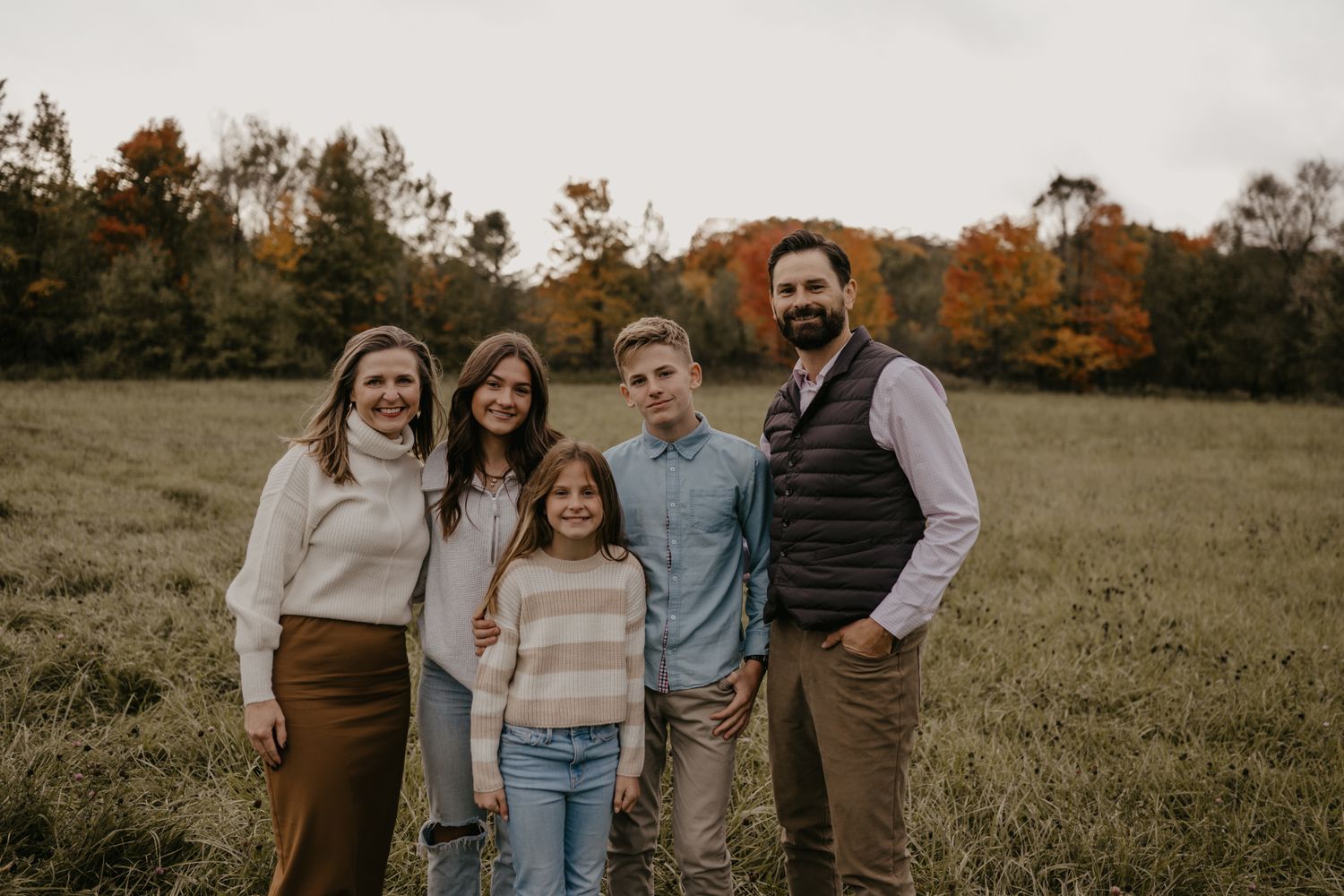 Family of five smiling together in an autumn field with colorful fall foliage trees in the background.