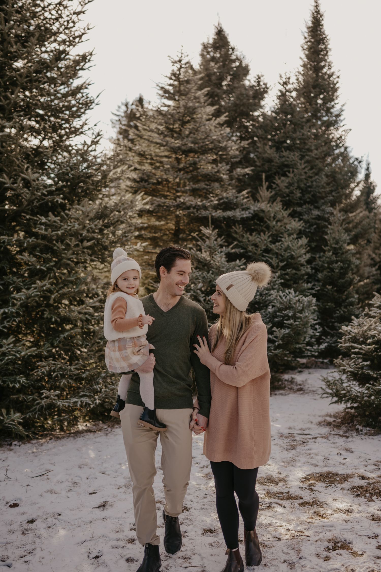 Family of three posing in snowy Christmas tree farm, parents in winter hats holding toddler, surrounded by pine trees.