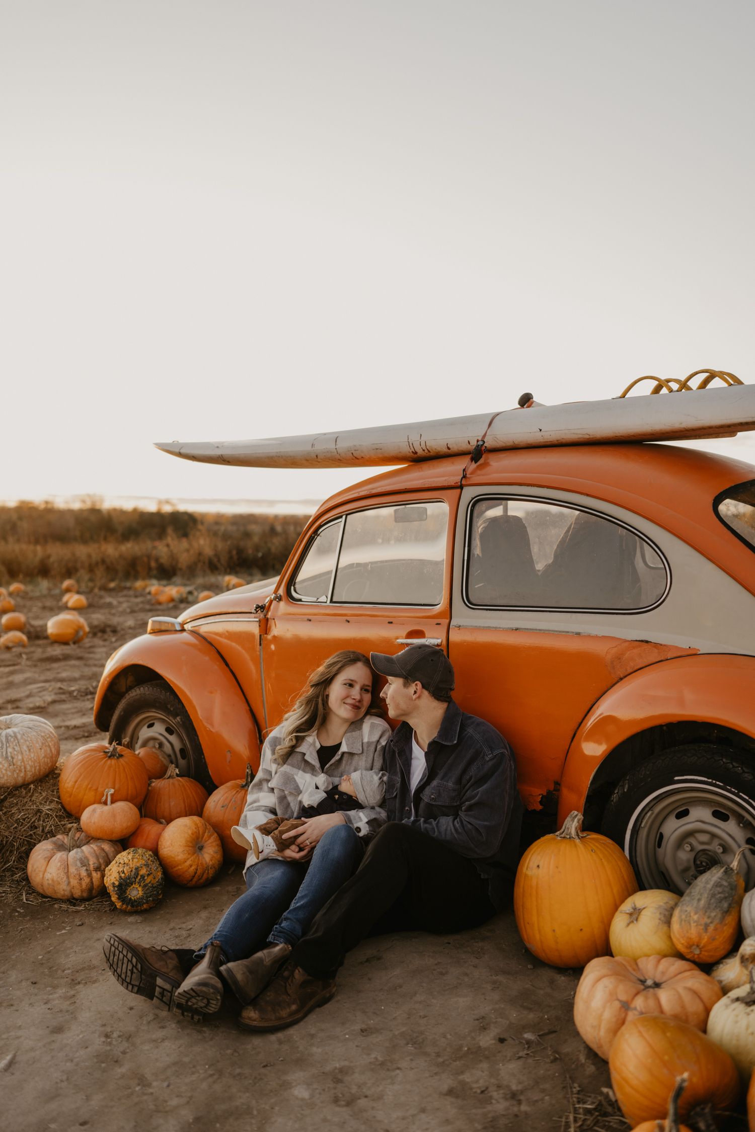 Couple sitting together by a vintage orange VW Beetle surrounded by pumpkins at a fall pumpkin patch.