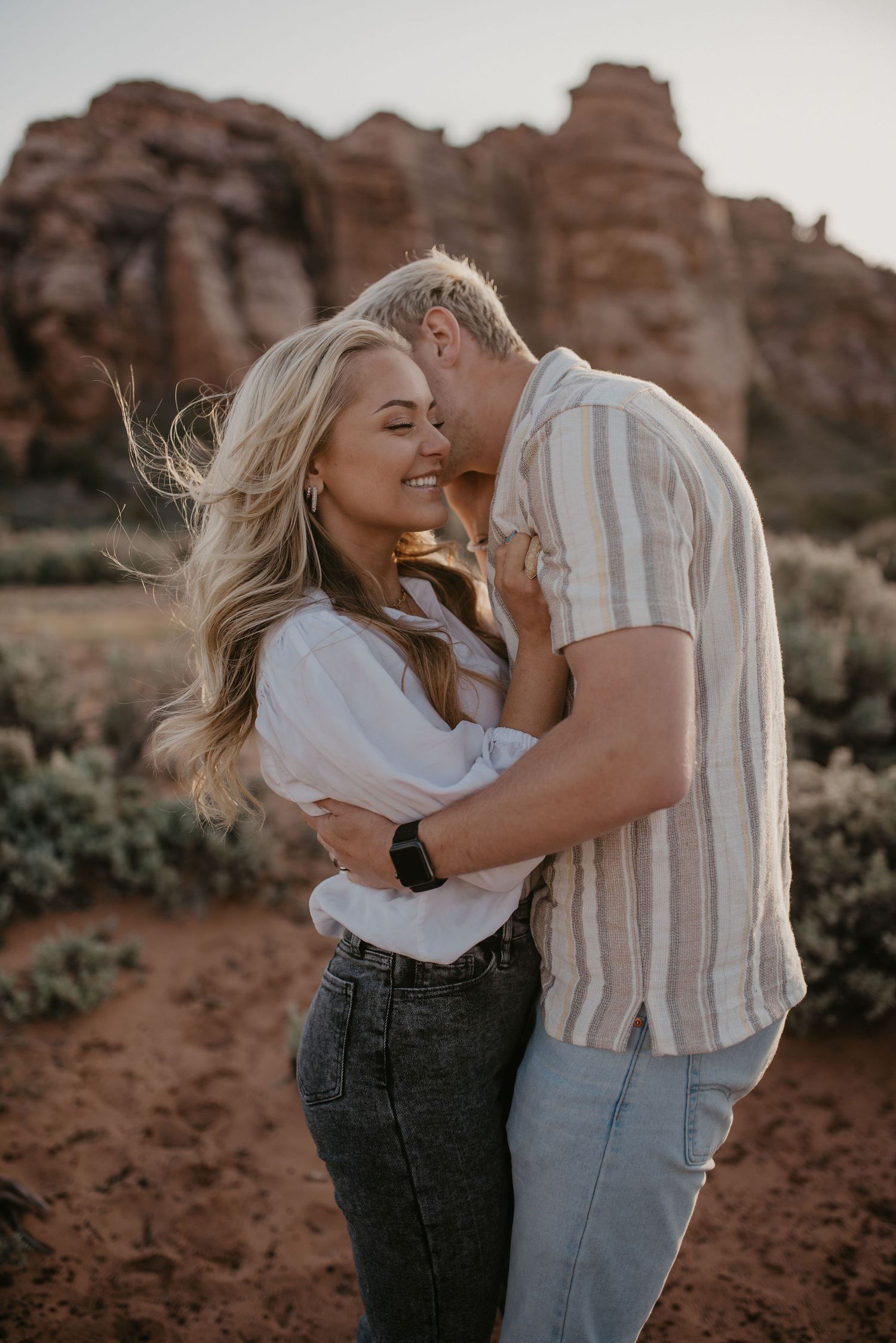 Couple embracing and smiling in a desert landscape with red rock formations at sunset.