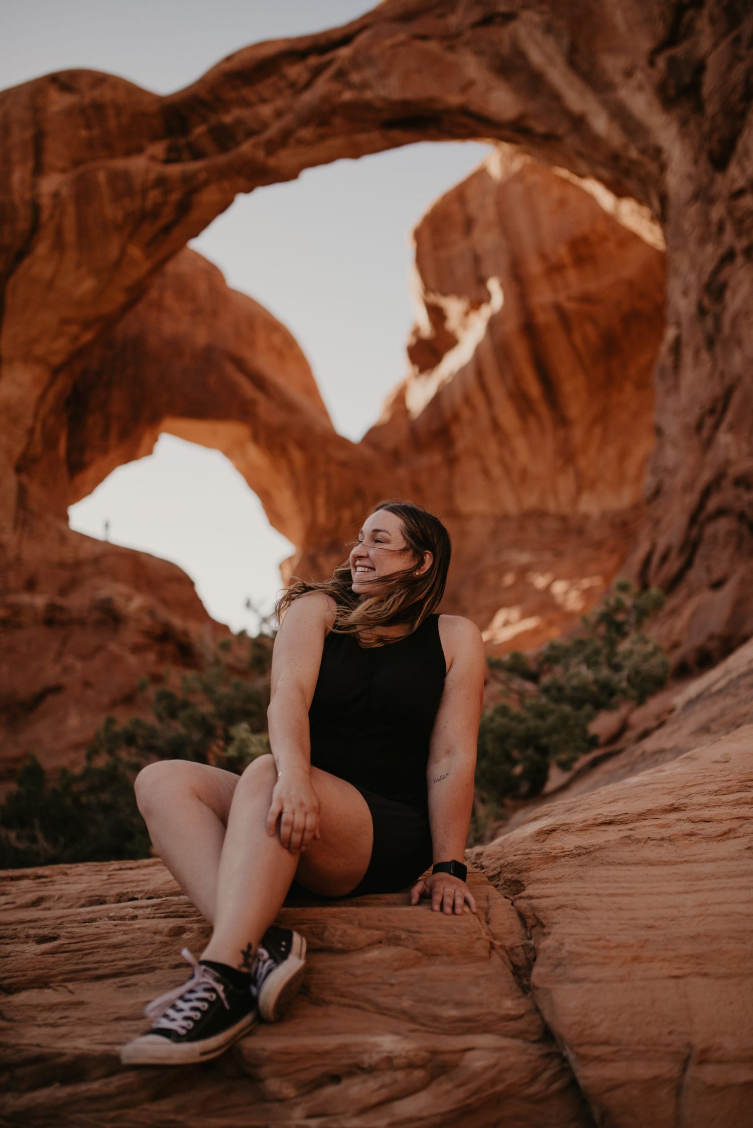 Smiling woman in black romper seated on red sandstone beneath the iconic Double Arch at Arches National Park.