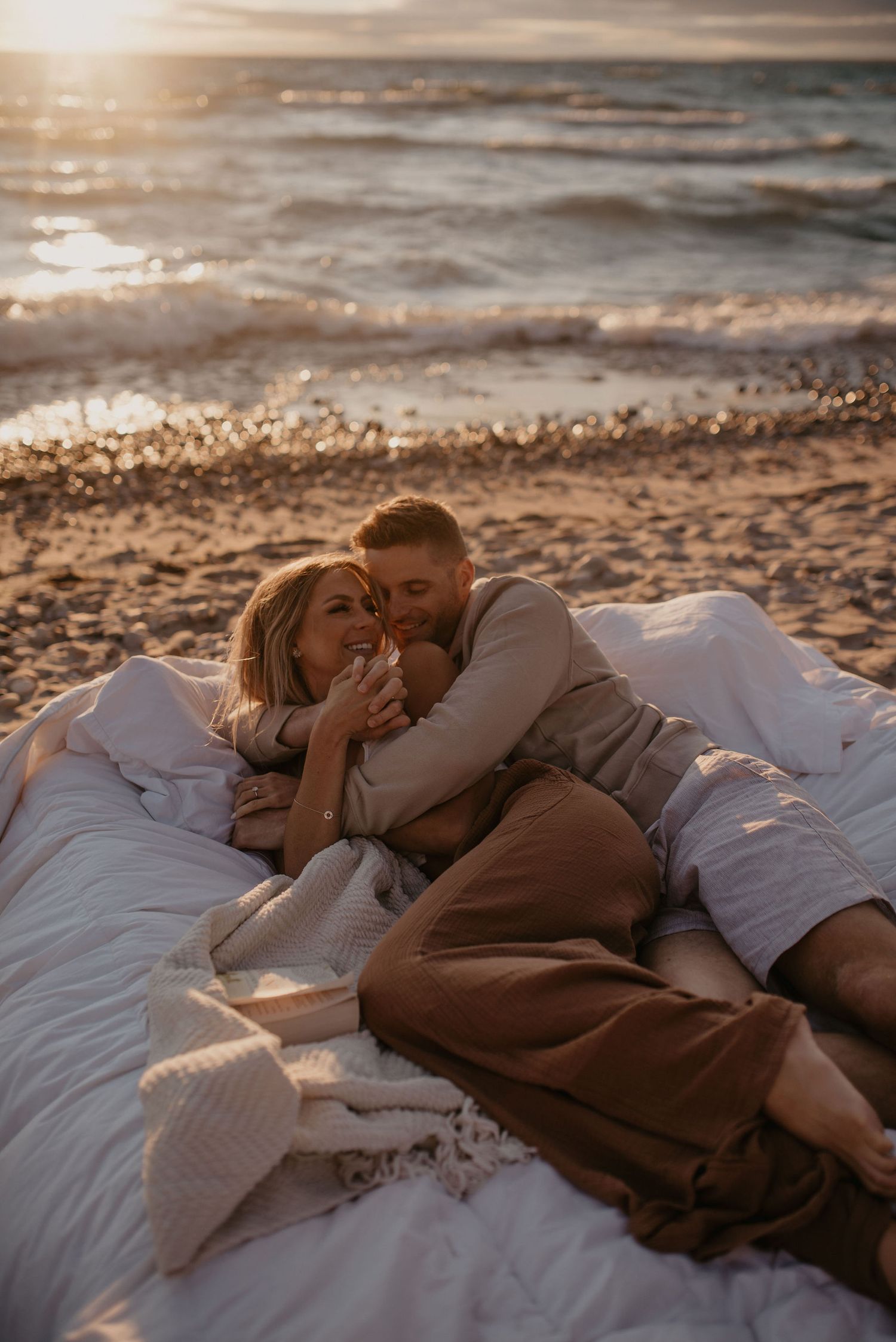 Romantic couple cuddling on white blankets on a sandy beach at sunset during a couples photo session.
