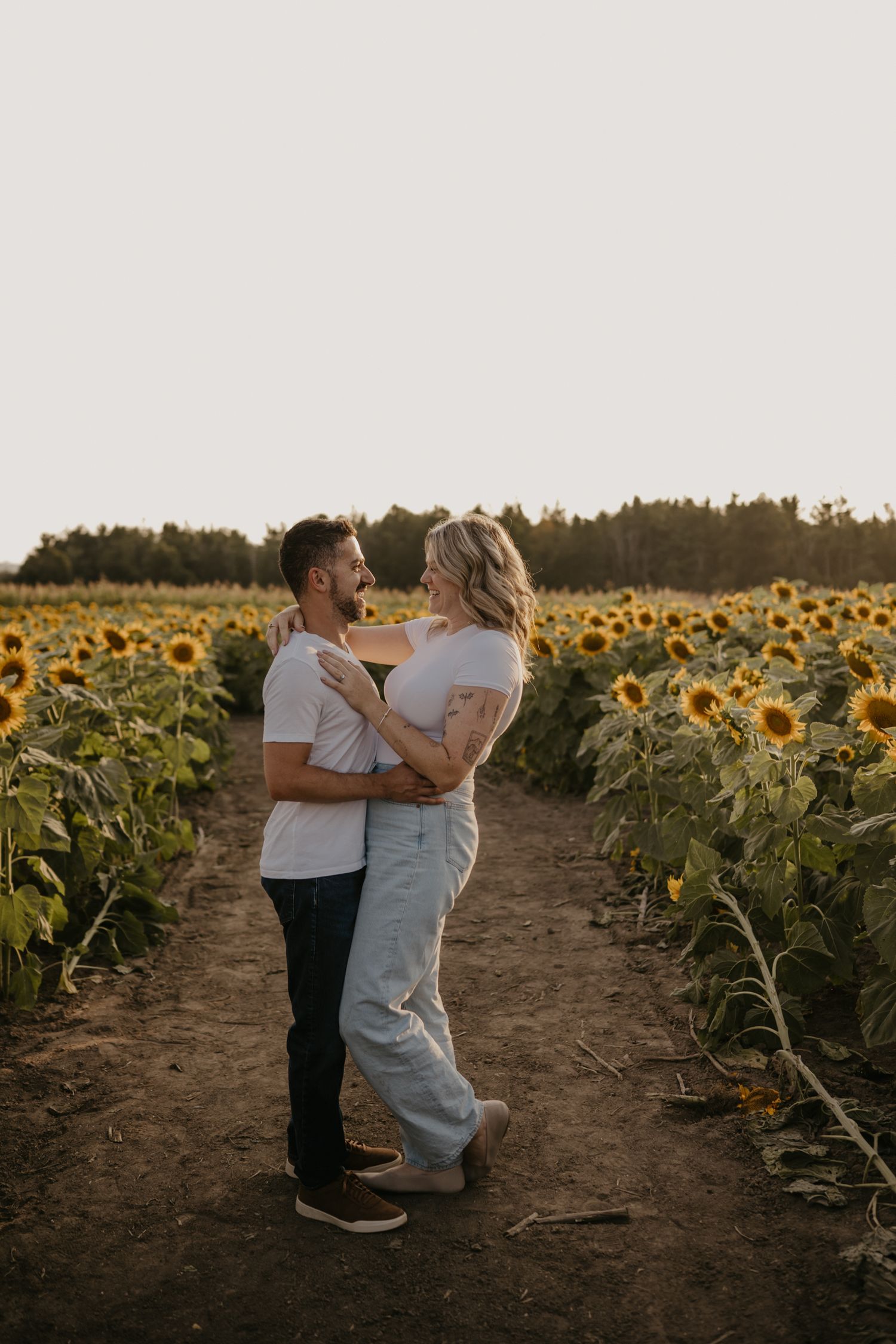 Couple embracing and smiling at each other in a sunflower field at golden hour.