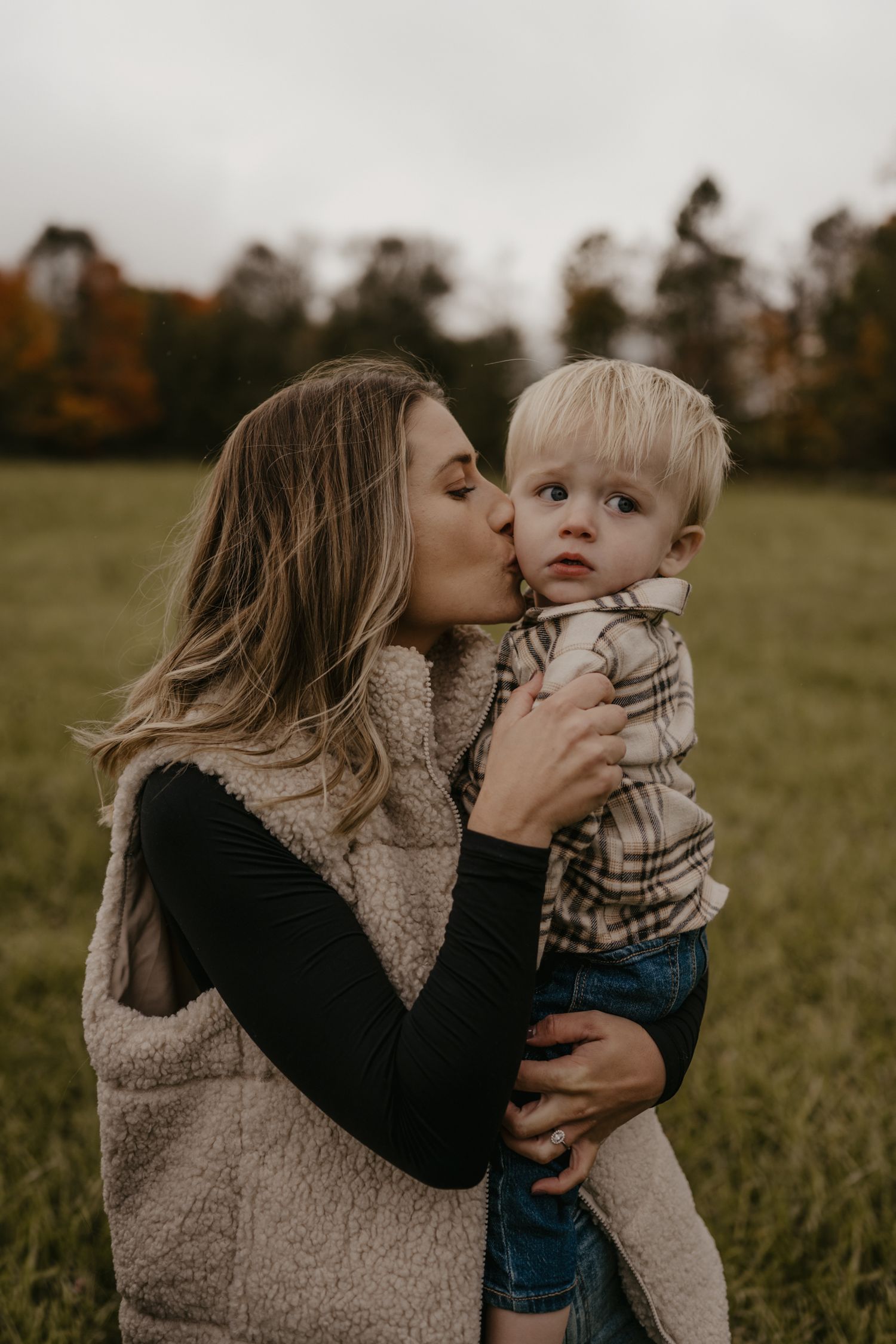 A mother kisses her toddler son on the cheek while holding him in an autumn field.
