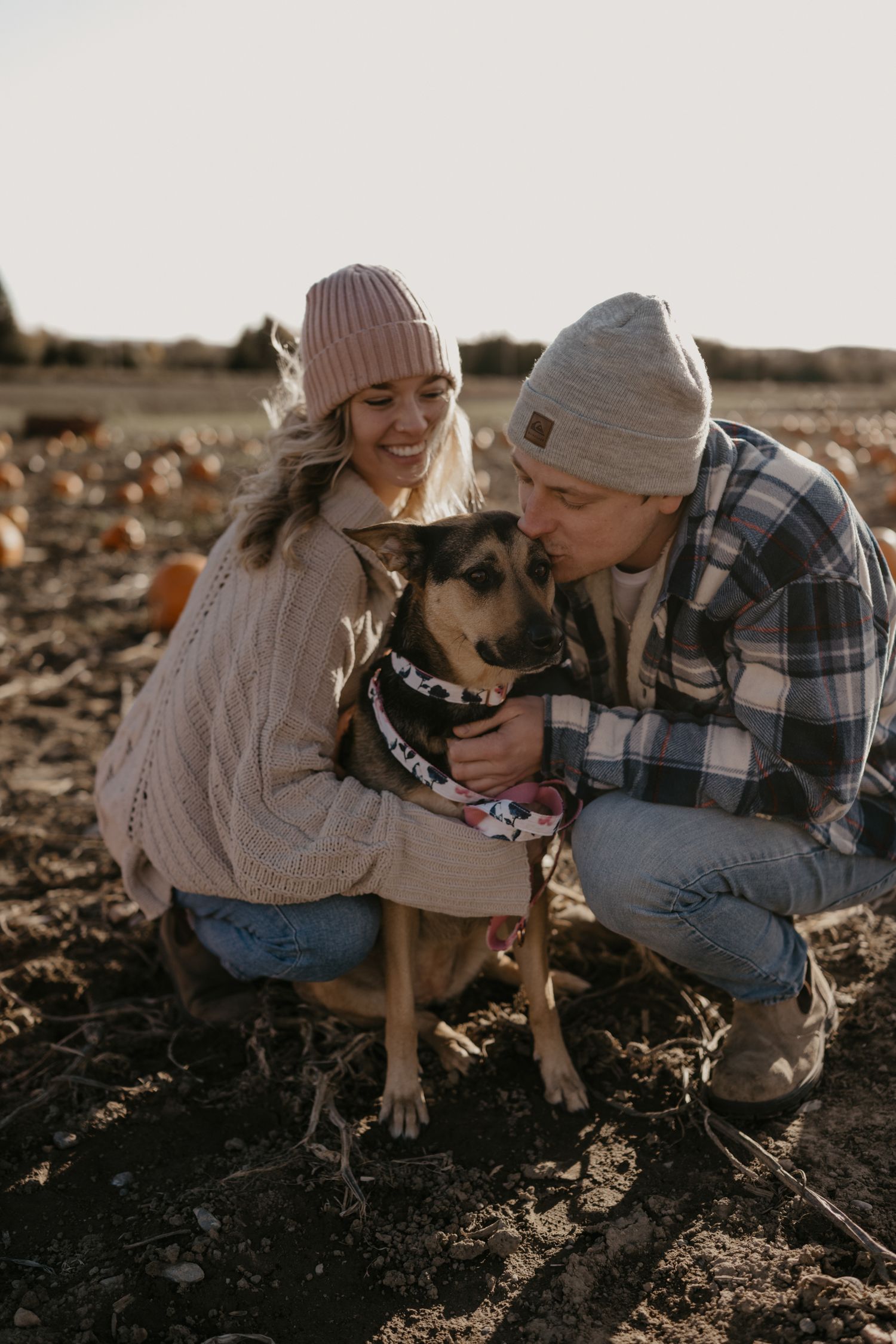 Couple in beanies and sweaters crouching with their dog in a pumpkin patch during golden hour.