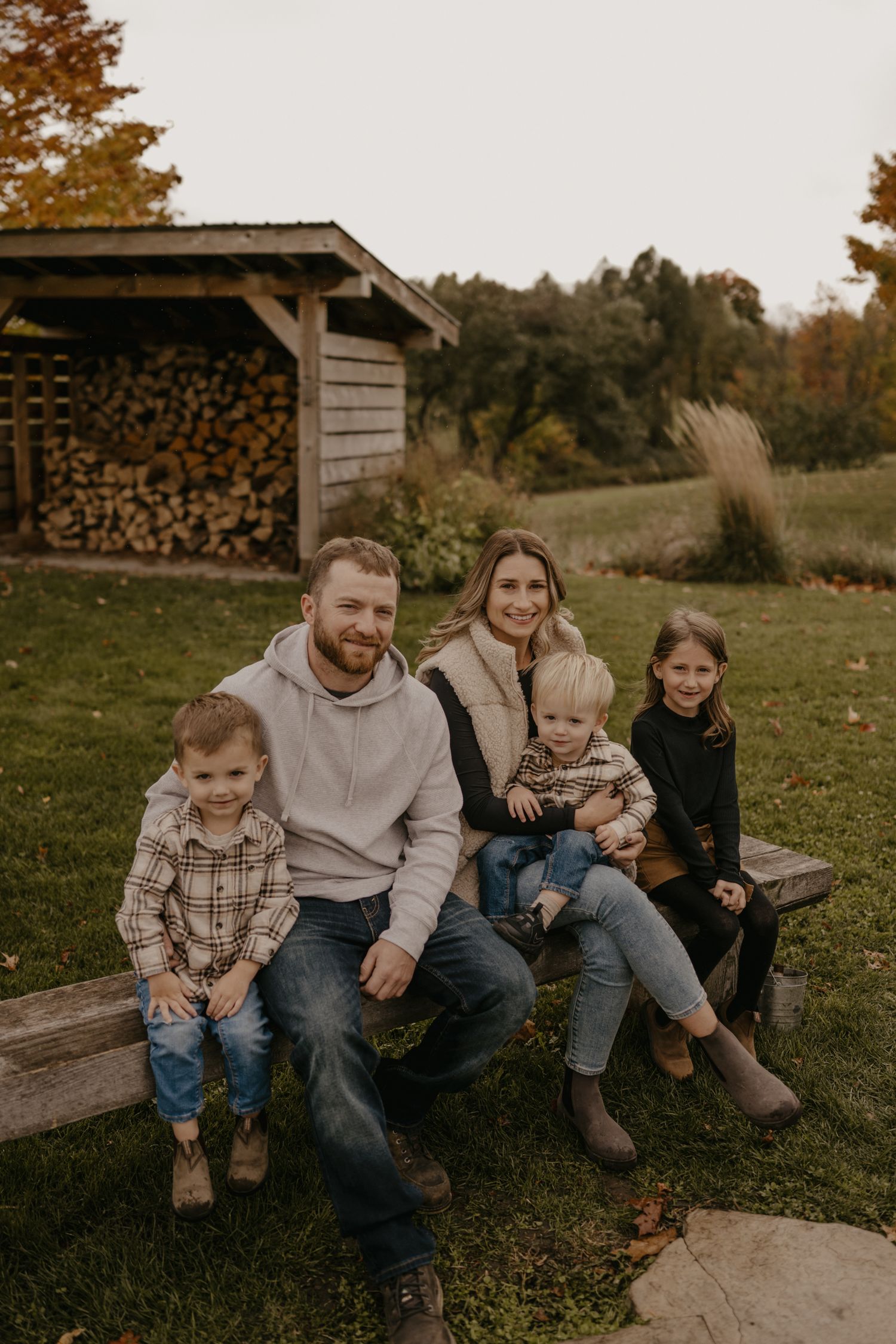 Family of five smiling together on a wooden bench outdoors in autumn, with a firewood shed and fall foliage in the background.
