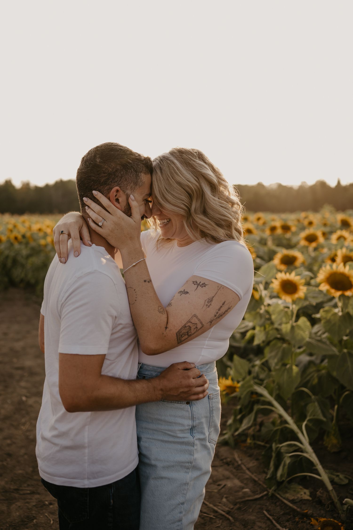 Couple with tattoos sharing an intimate forehead touch in a sunflower field during warm sunset.