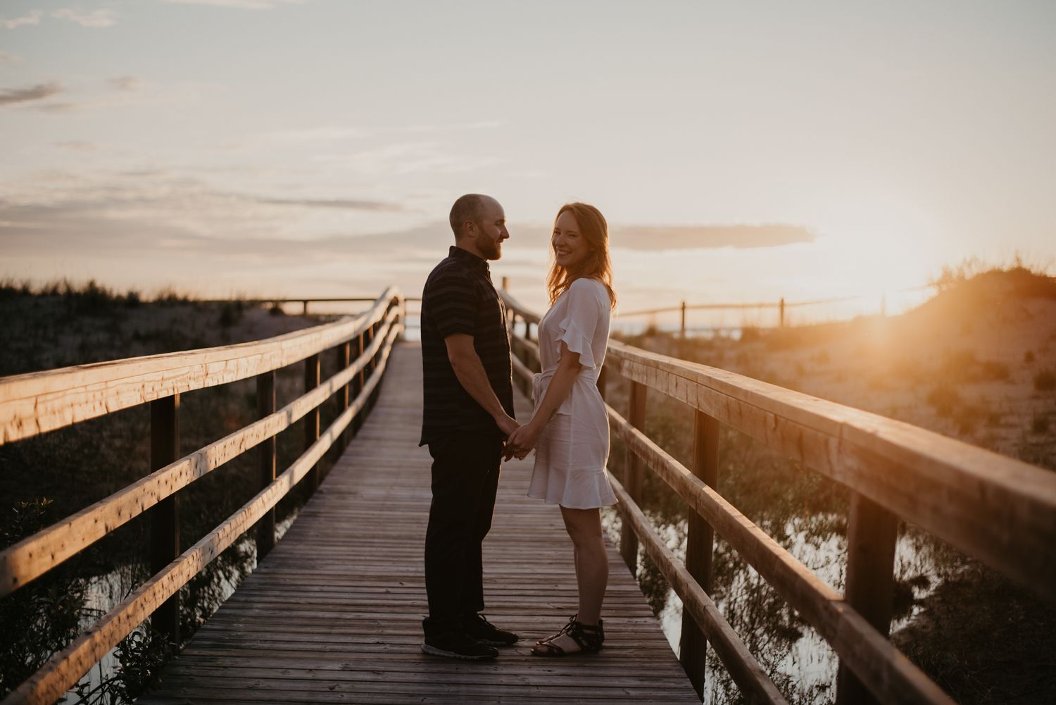 Couple holding hands facing each other on a wooden boardwalk during a golden sunset at the beach.