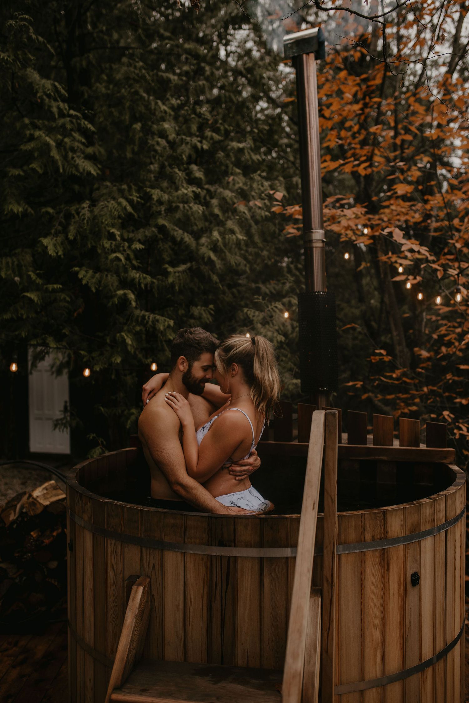Couple embracing intimately in a wooden hot tub surrounded by autumn forest lights.