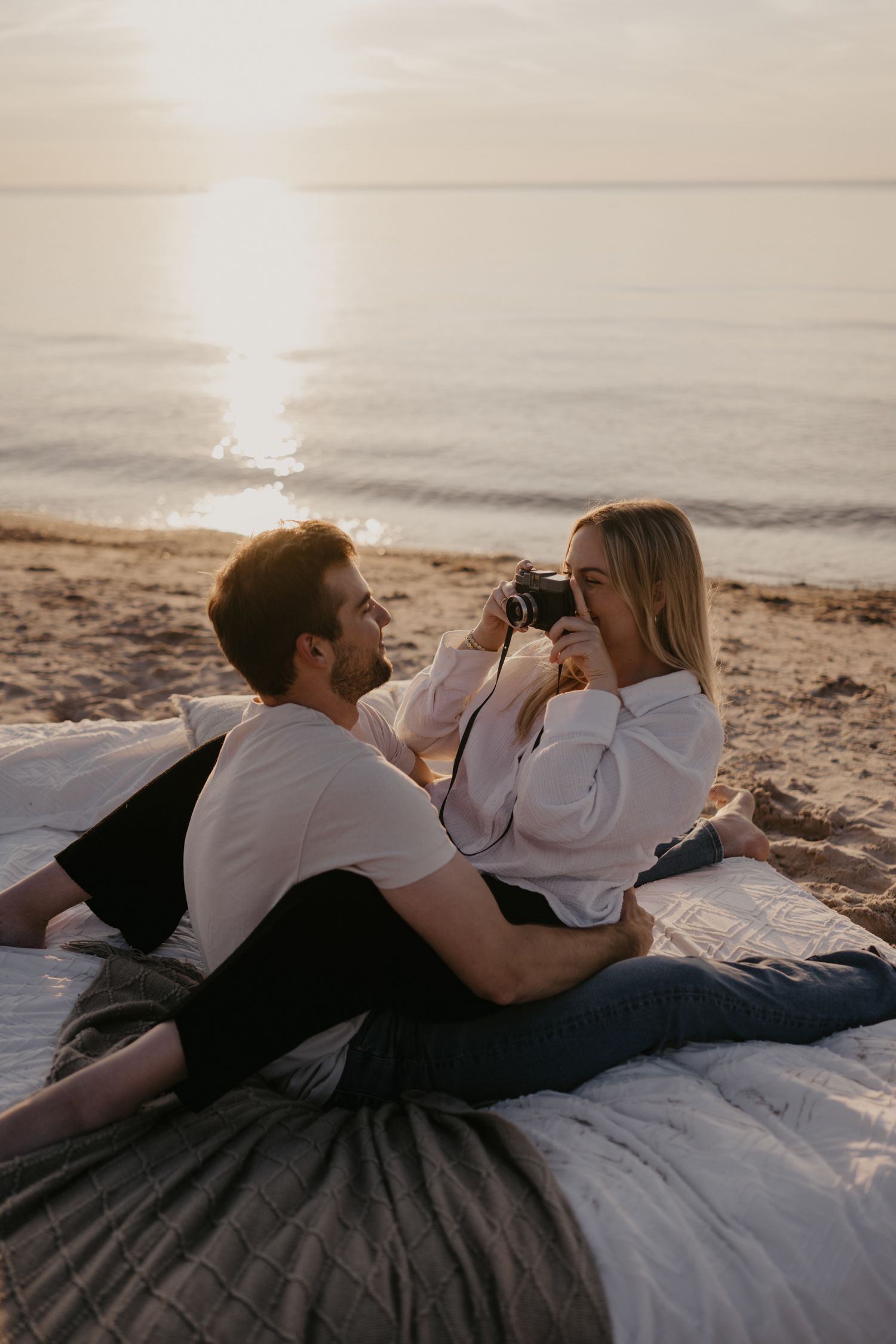 Couple relaxing on beach blanket at sunset, woman photographing man with vintage camera.