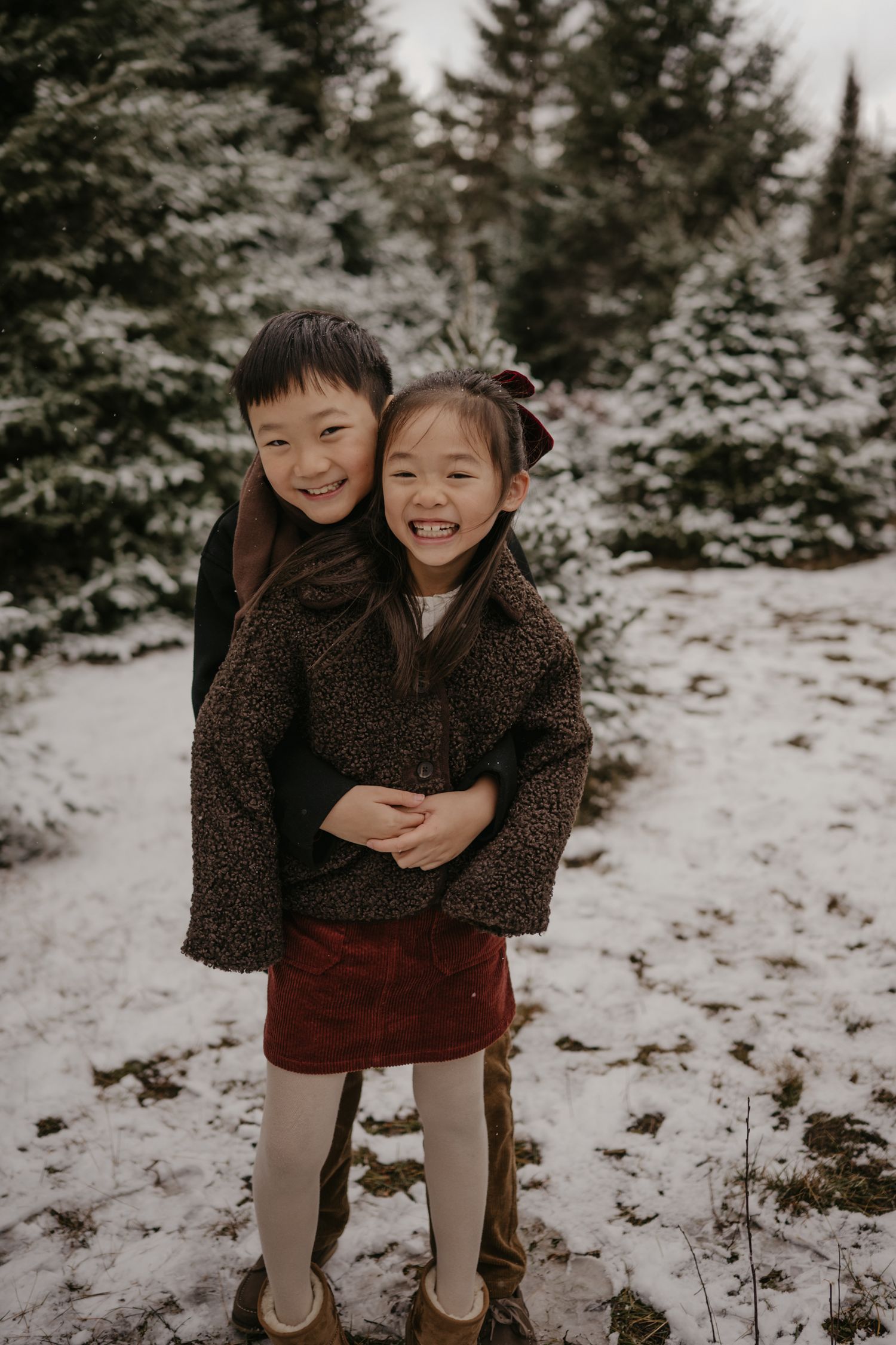 Two smiling Asian children hugging in a snowy evergreen forest, wearing cozy winter outfits.