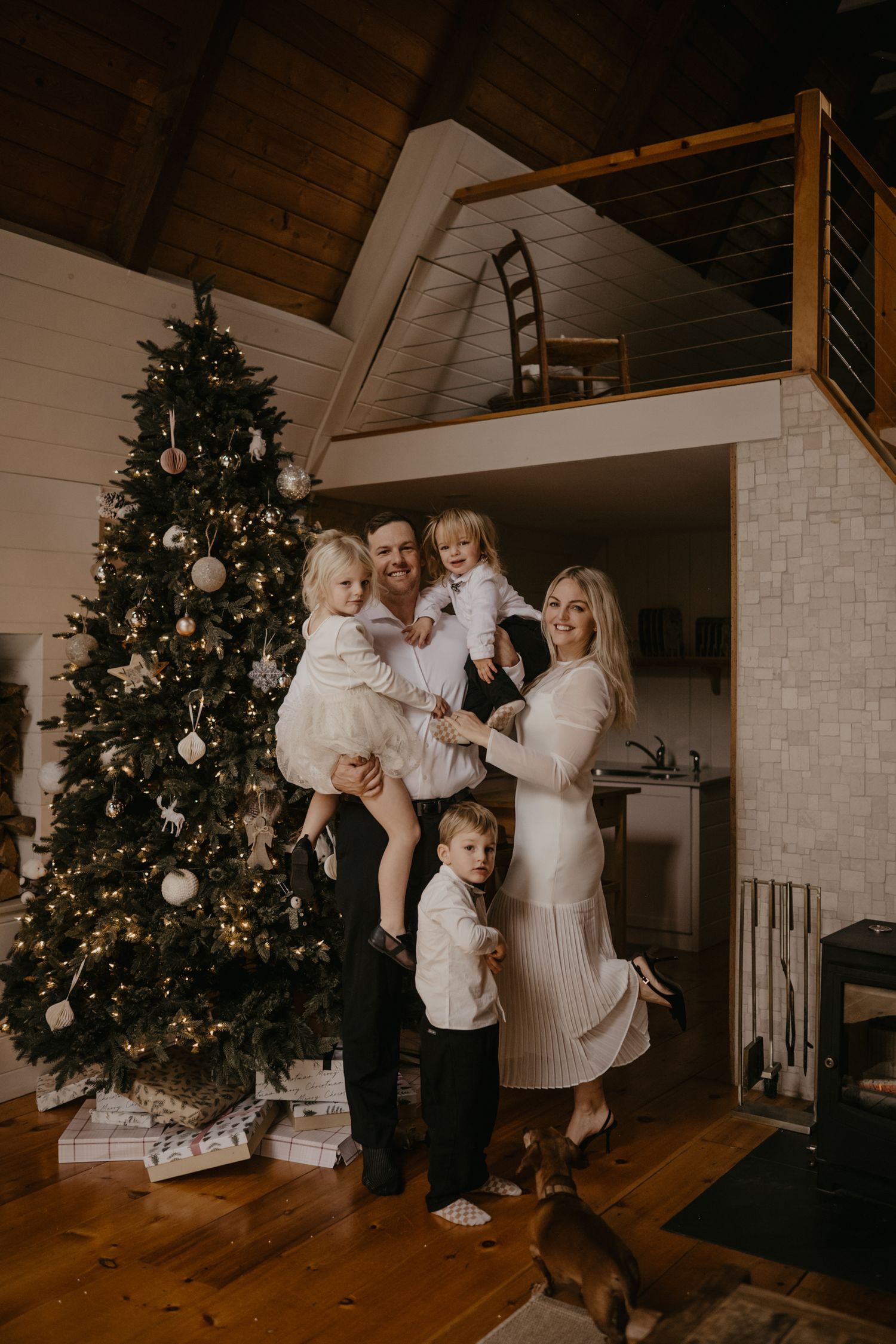 Smiling family of five posing beside a tall decorated Christmas tree inside a cozy A-frame cabin with loft.