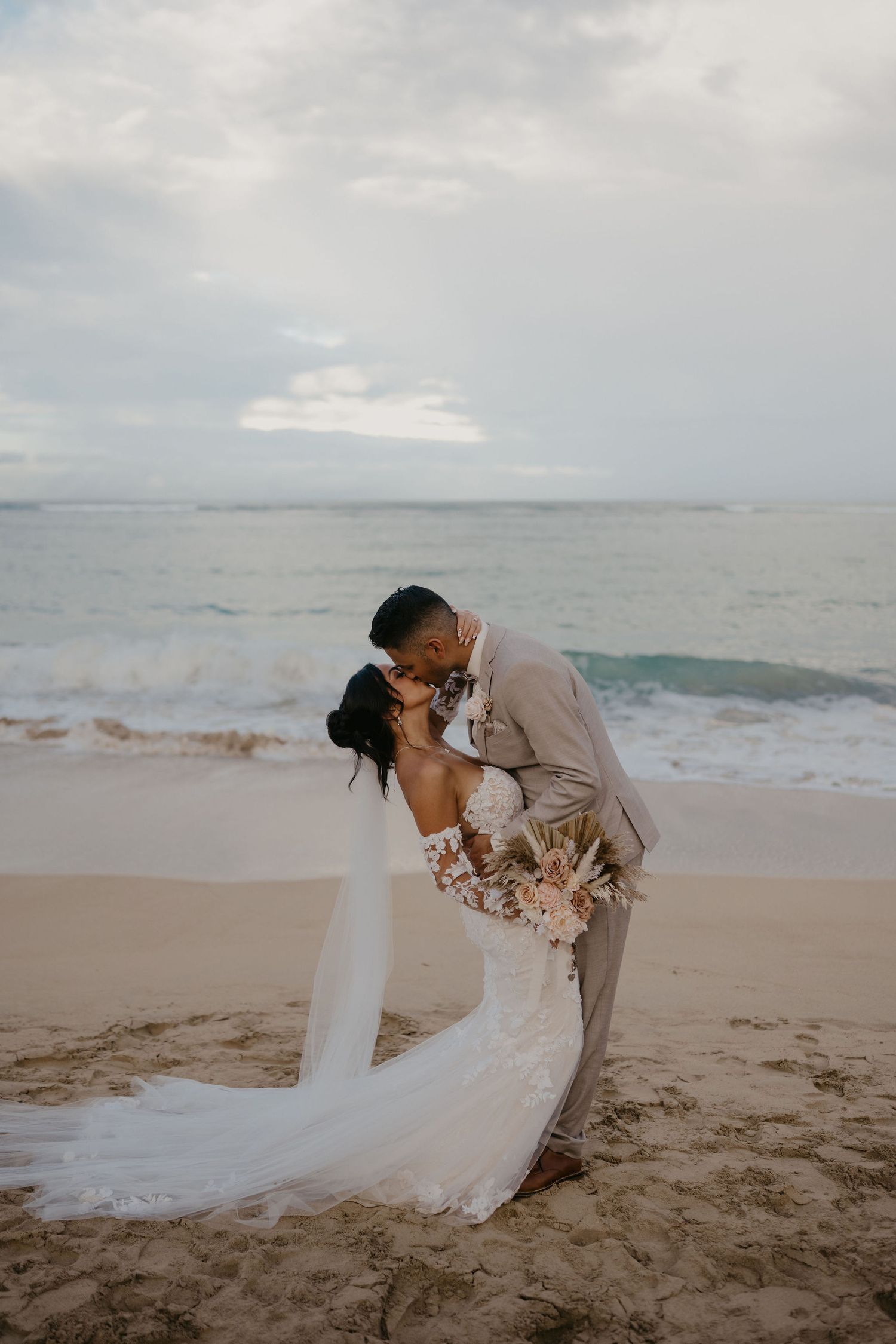 Bride and groom sharing a romantic dip kiss on sandy beach with ocean waves behind them.
