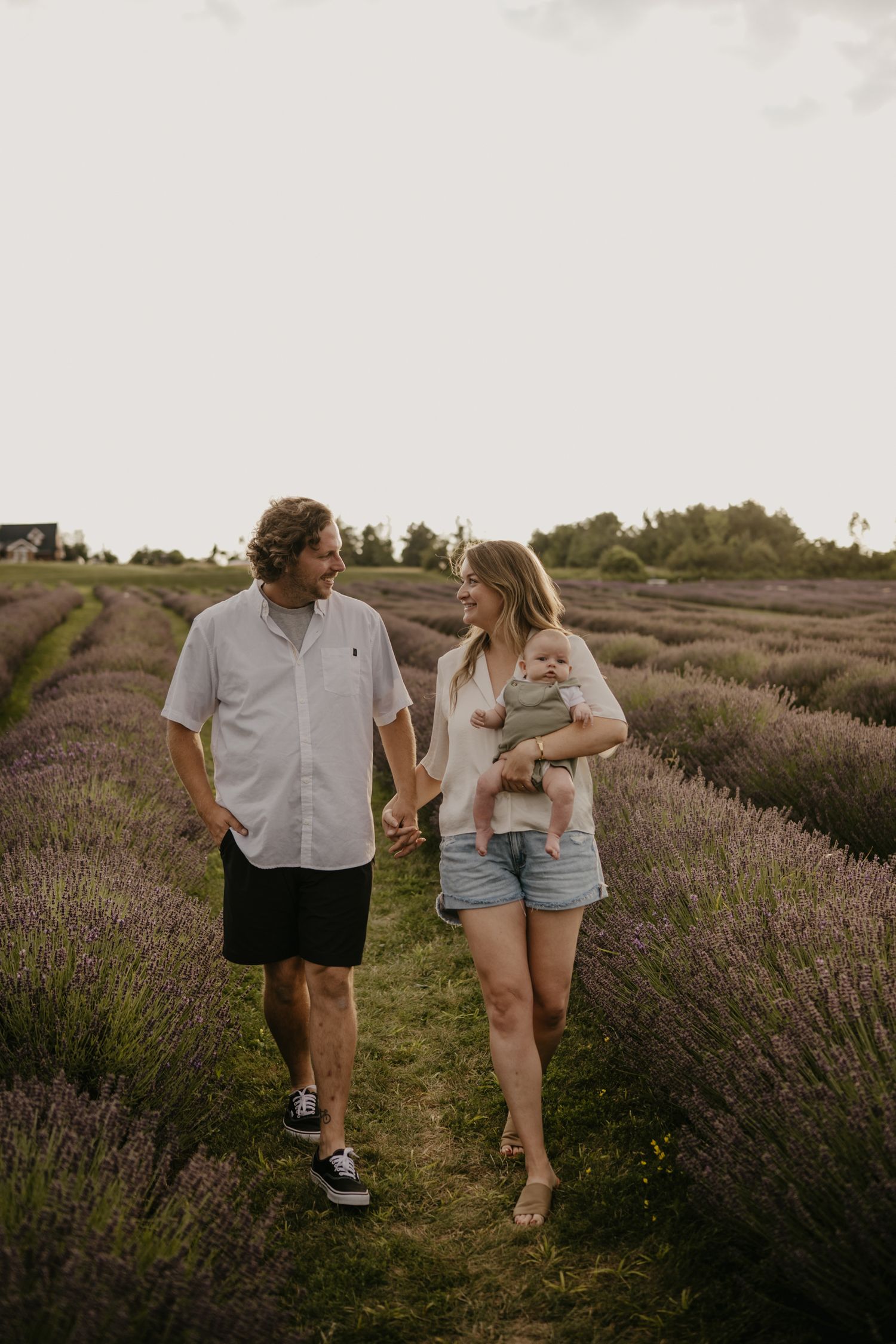 Smiling couple holding hands and carrying newborn baby walking through lavender field.