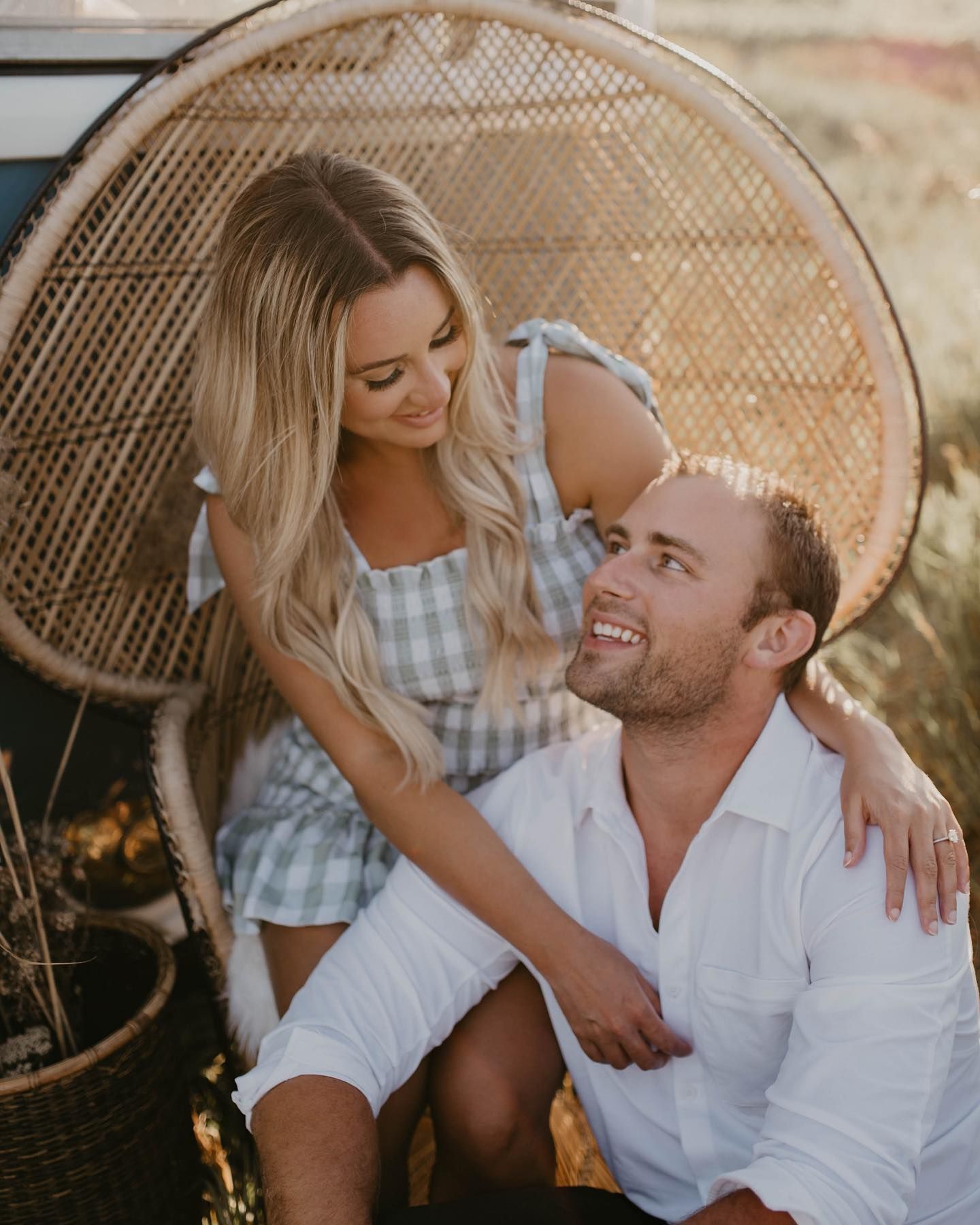 Smiling blonde couple posing together in a peacock chair during golden hour outdoor engagement session.