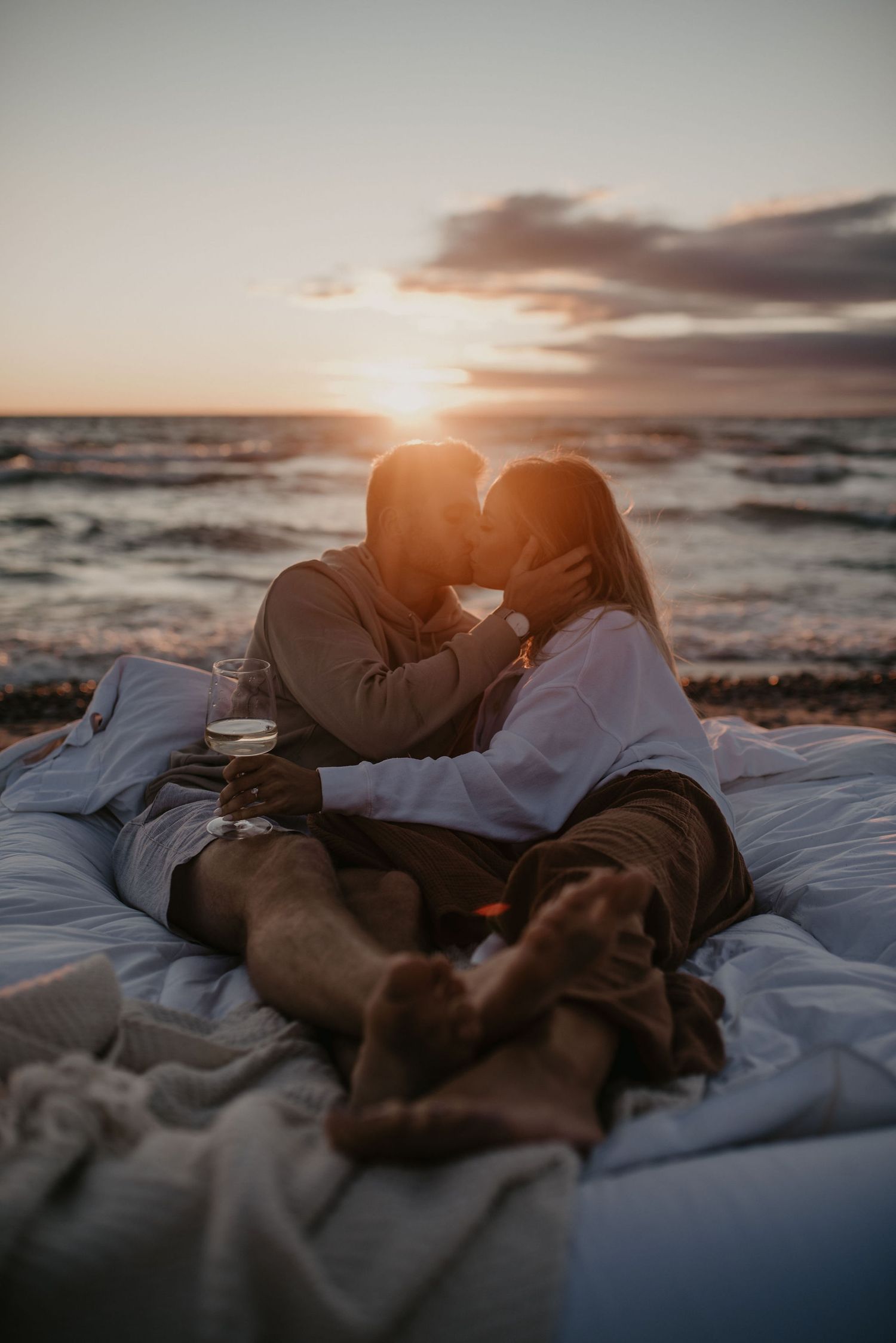Couple sharing a romantic kiss on a white blanket at the beach during a vibrant sunset.