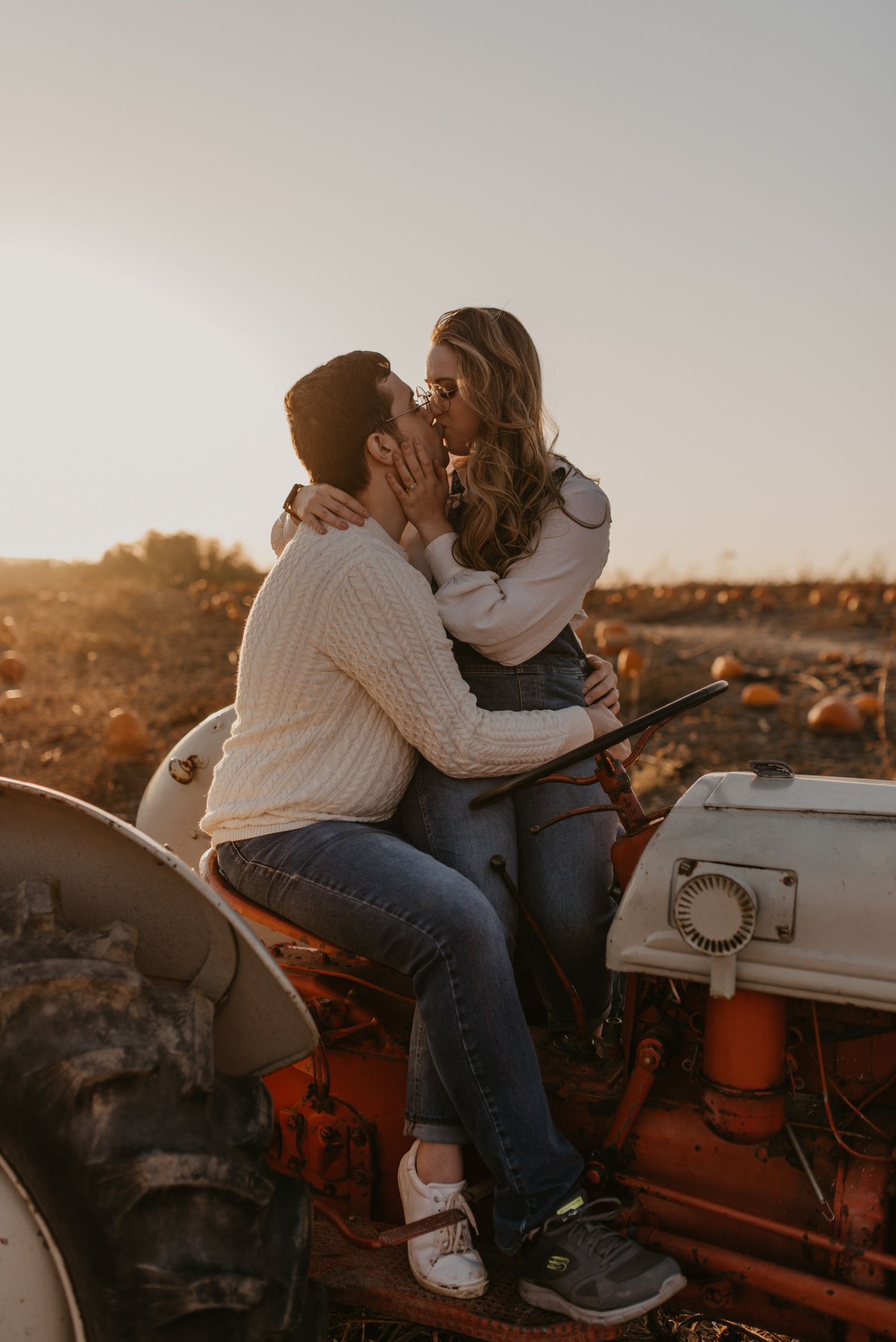 Couple sharing a romantic kiss on vintage red tractor in a pumpkin patch at golden hour.