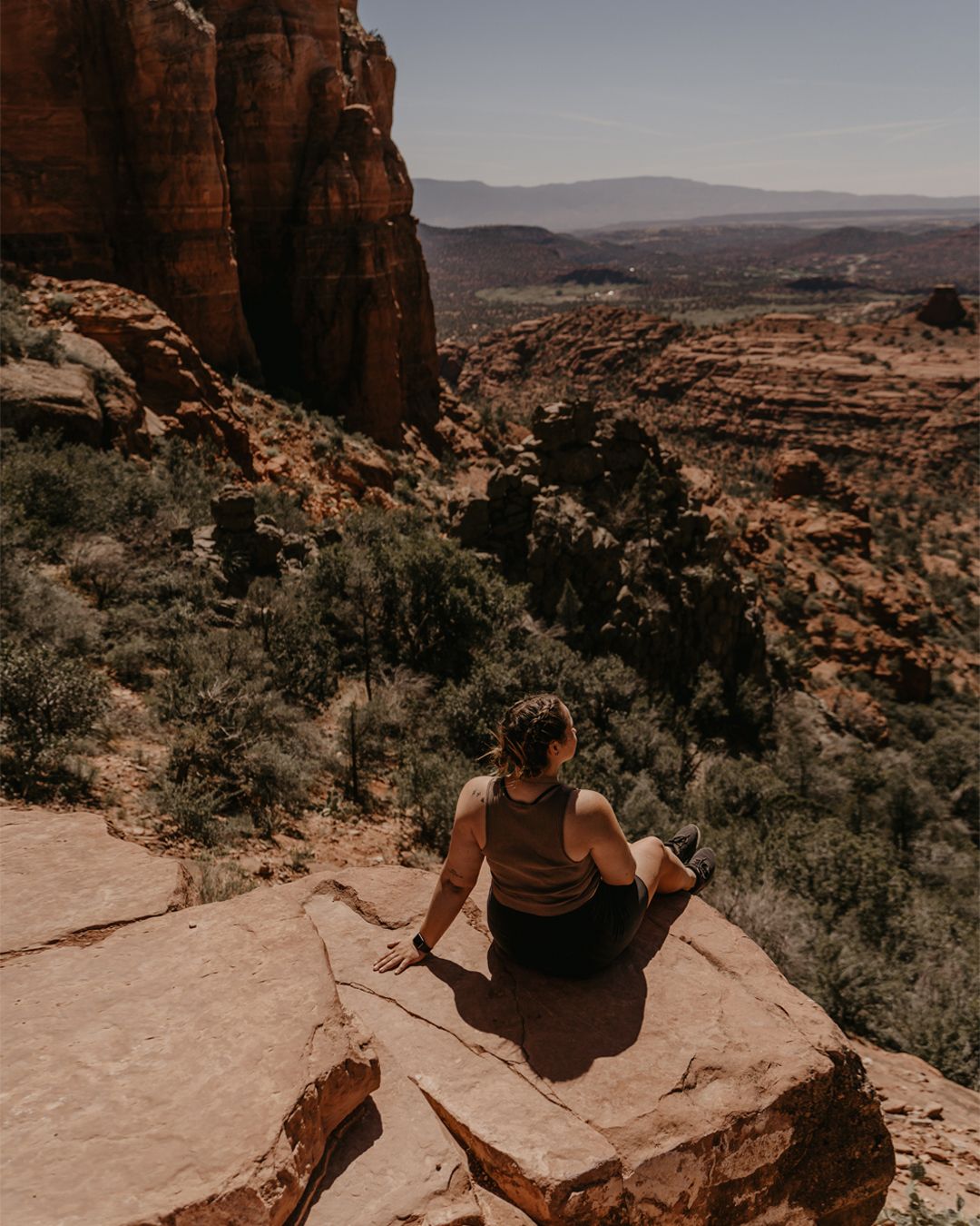 Person sitting on red rock ledge overlooking a vast canyon landscape with green shrubs and blue sky.