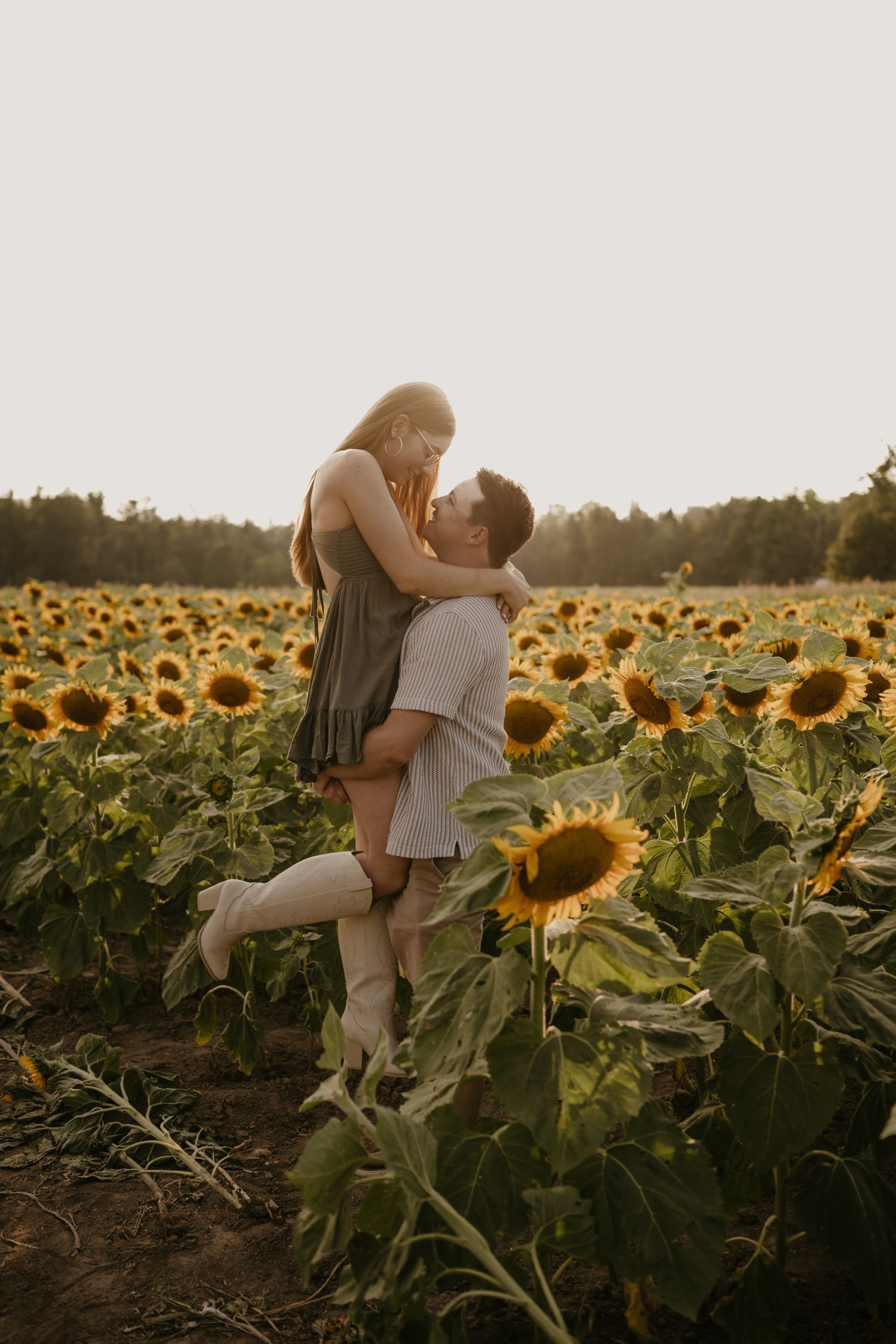Man lifting woman in green dress for a kiss among tall sunflowers during golden hour sunset.