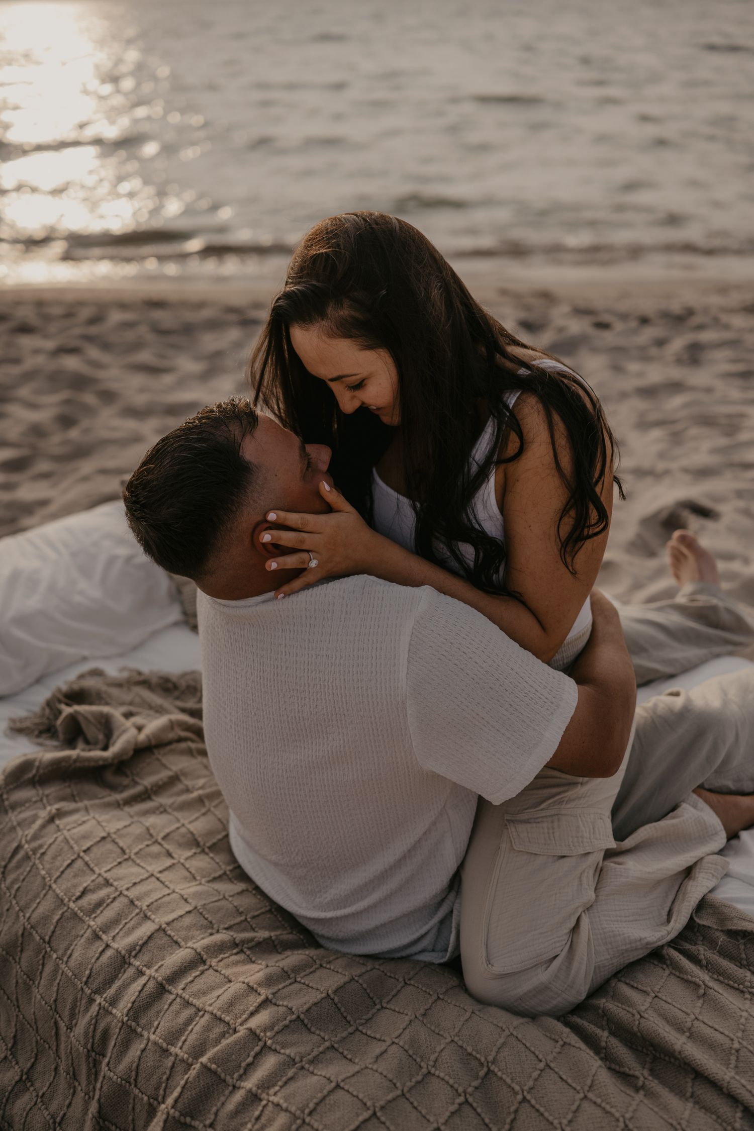 Couple embracing on beach blanket at sunset, woman sitting on man's lap laughing joyfully.