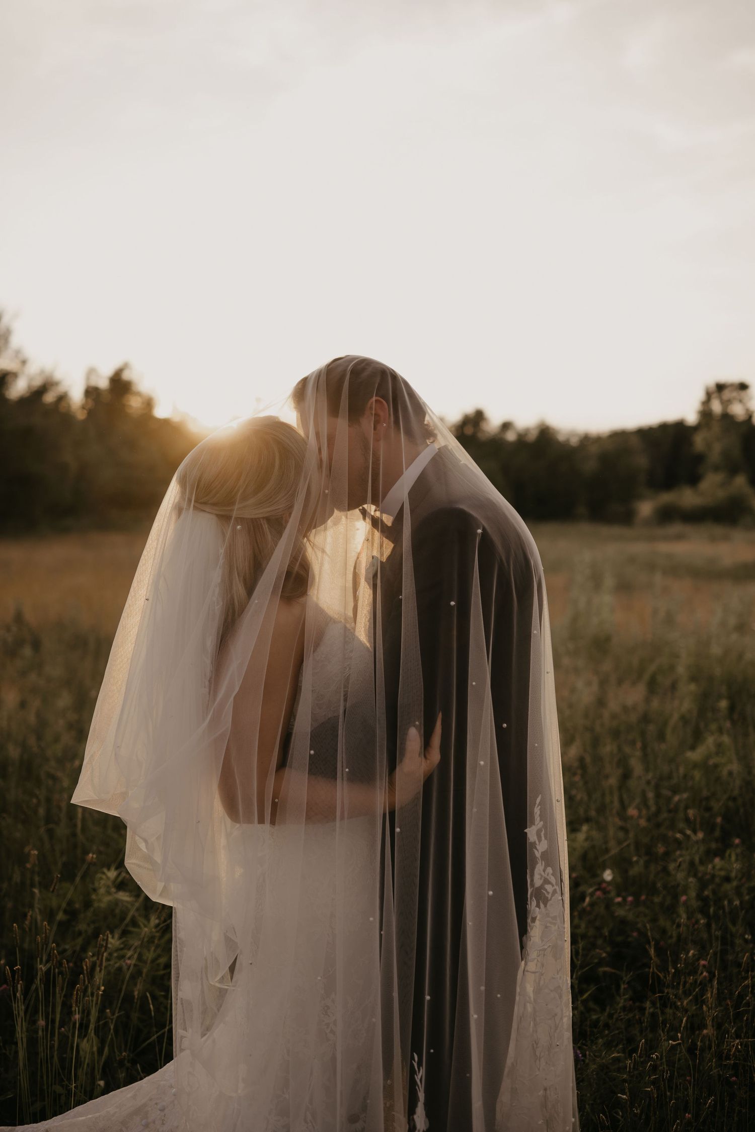 Bride and groom sharing a romantic kiss under a flowing veil at golden hour in a lush green field.
