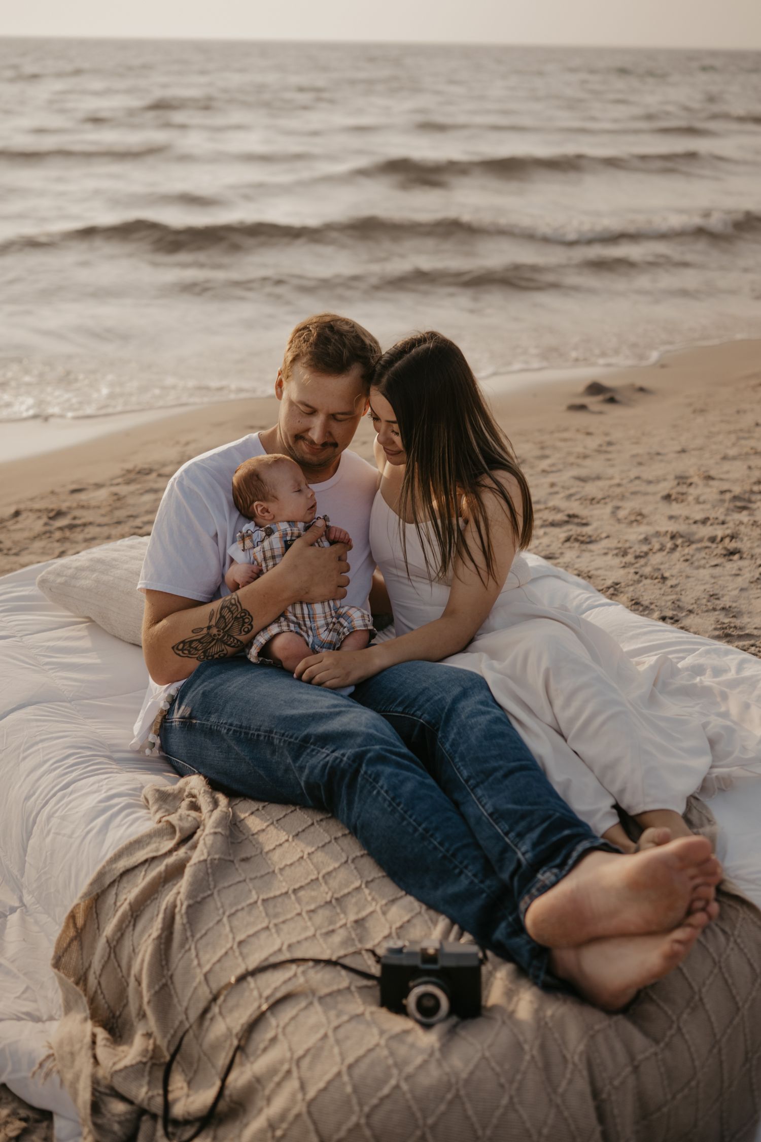Parents sitting on beach blanket with newborn baby, touching foreheads near ocean waves.