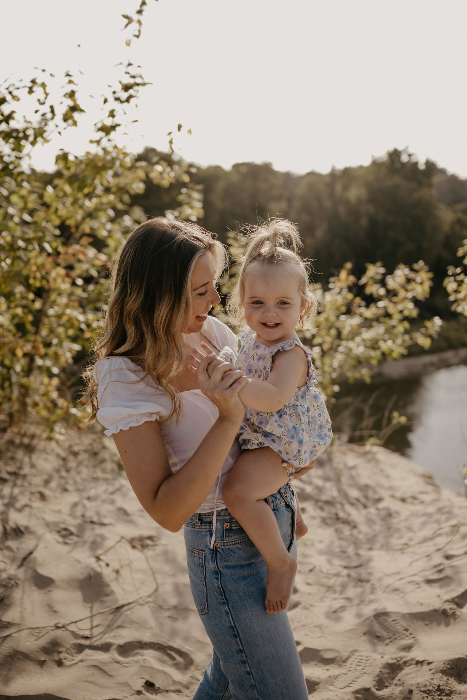 Smiling mother holding laughing toddler girl on sandy dunes with golden sunlight and greenery behind.