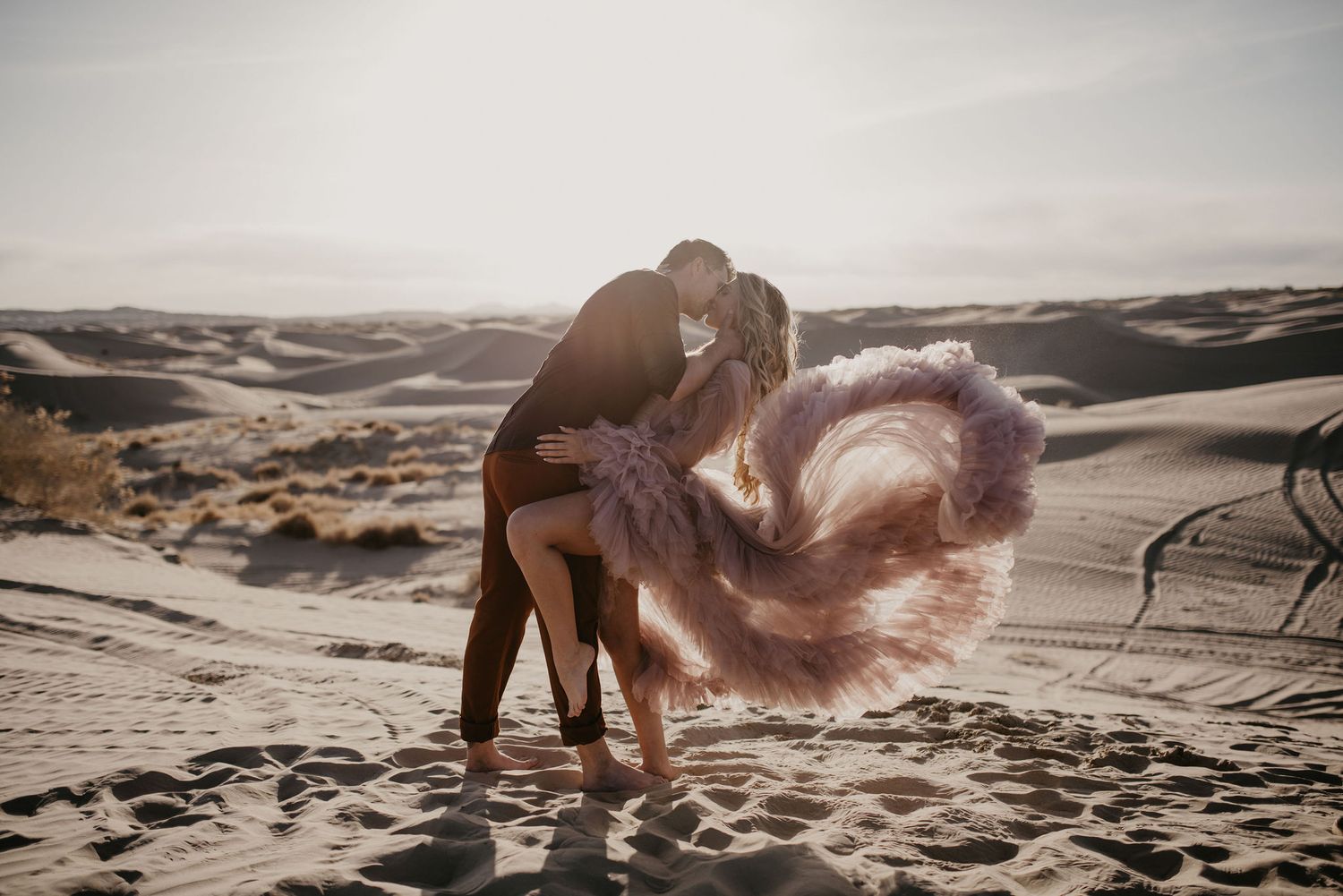 Couple sharing a romantic kiss on sand dunes with a flowing pink tulle dress.