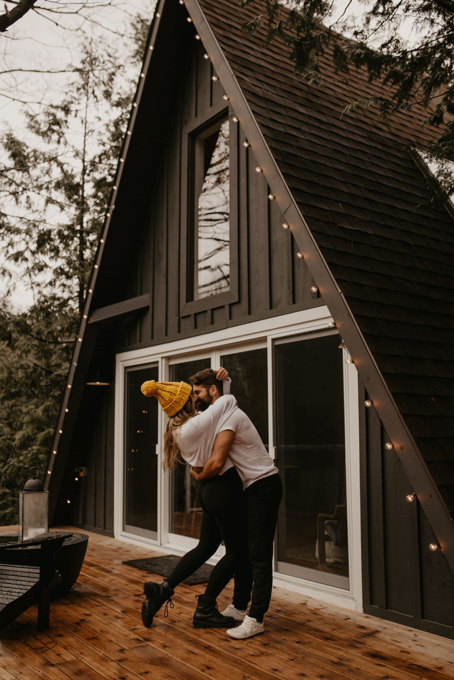 Couple embracing and dancing on wooden deck outside a dark A-frame cabin with string lights in the forest.