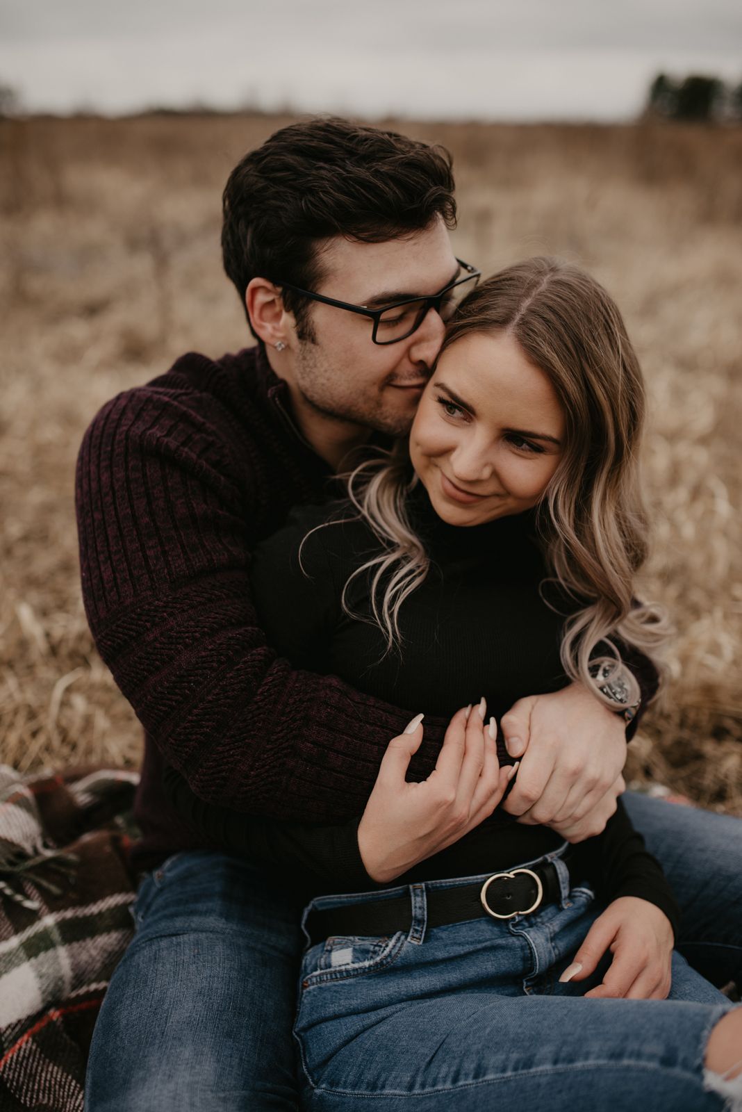 Young couple snuggling on a plaid blanket in a dry autumn field during a cozy engagement session.