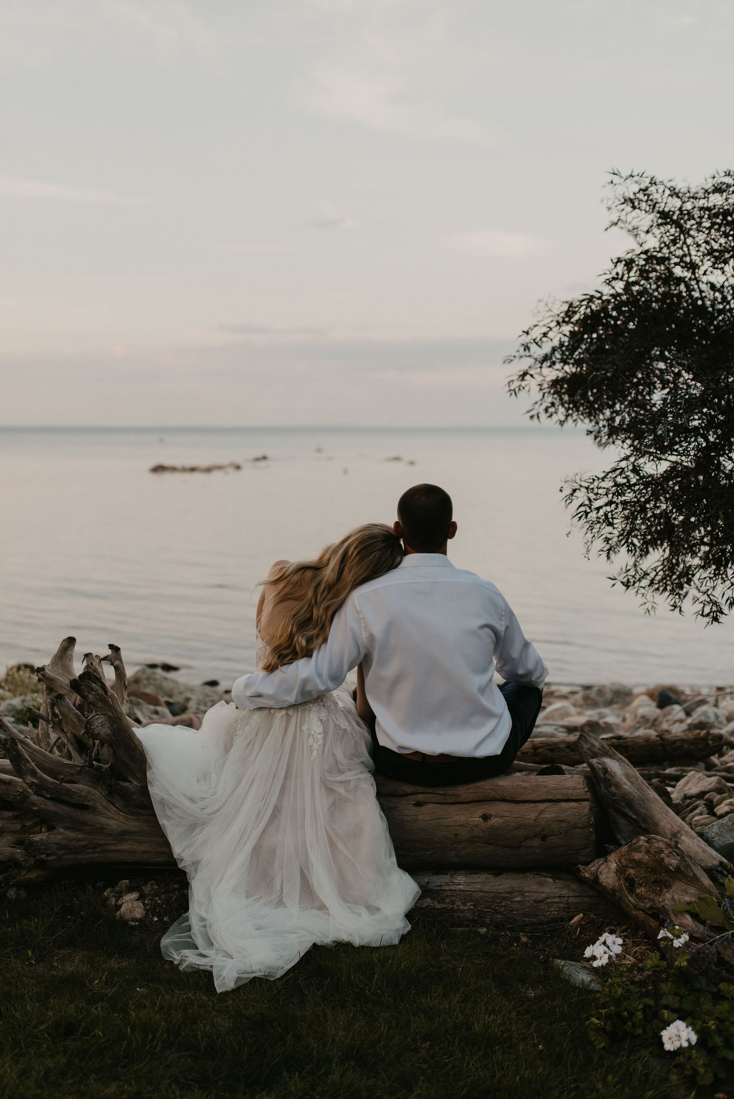 Bride and groom sit together on driftwood log gazing at calm ocean at dusk.
